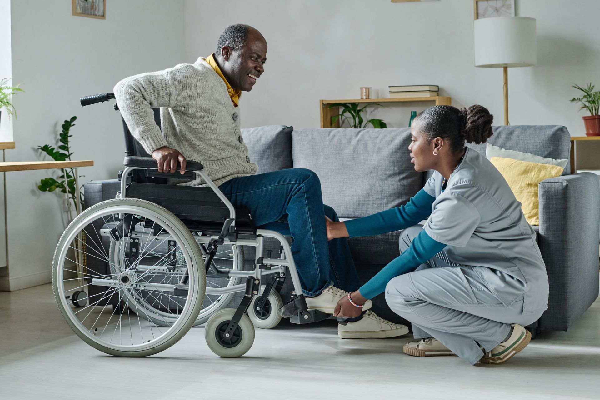Caregiver kneeling to help a person in a wheelchair adjust their shoe in a living room