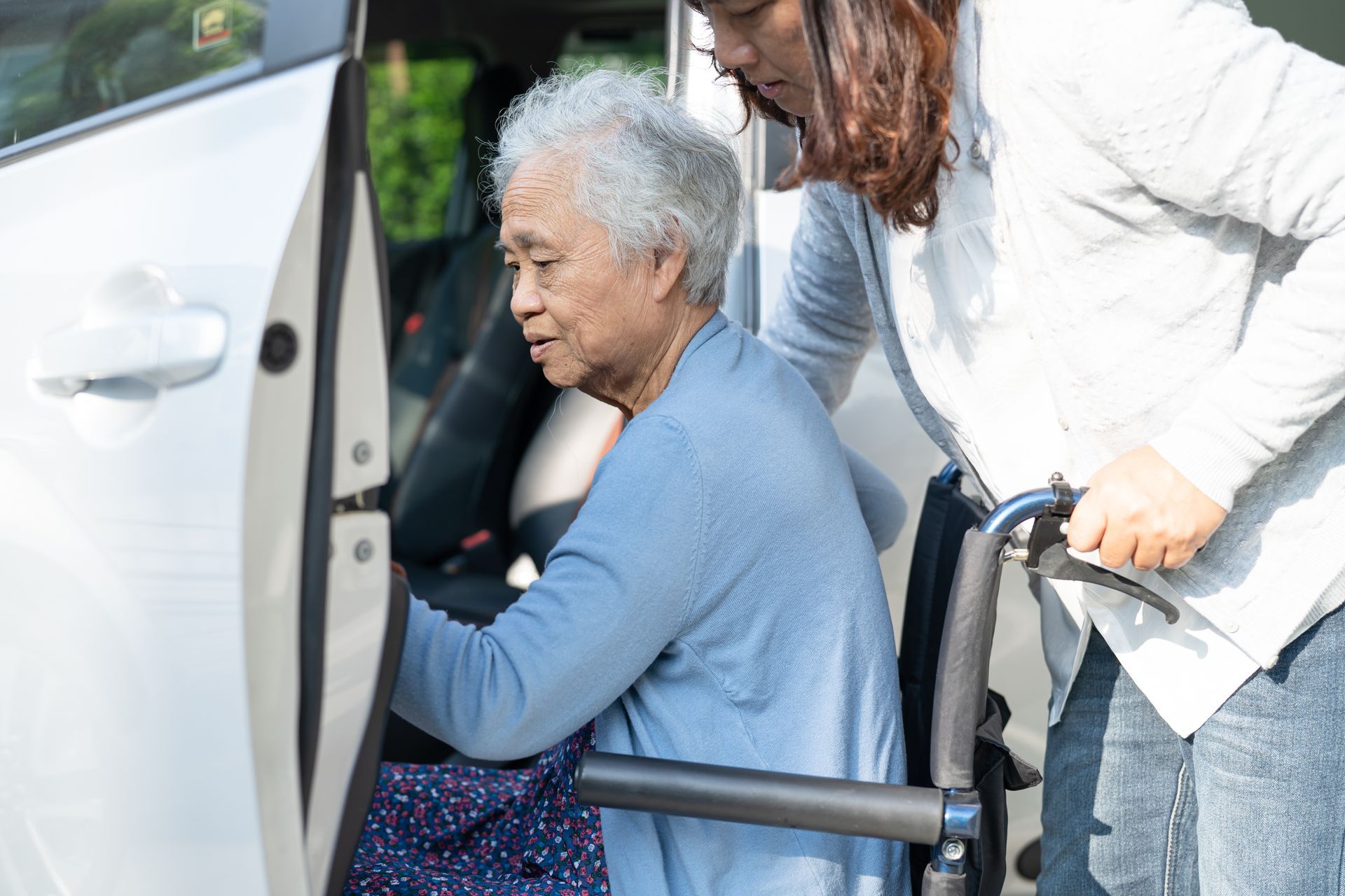 Woman helping an elderly passenger into a car from a wheelchair outdoors