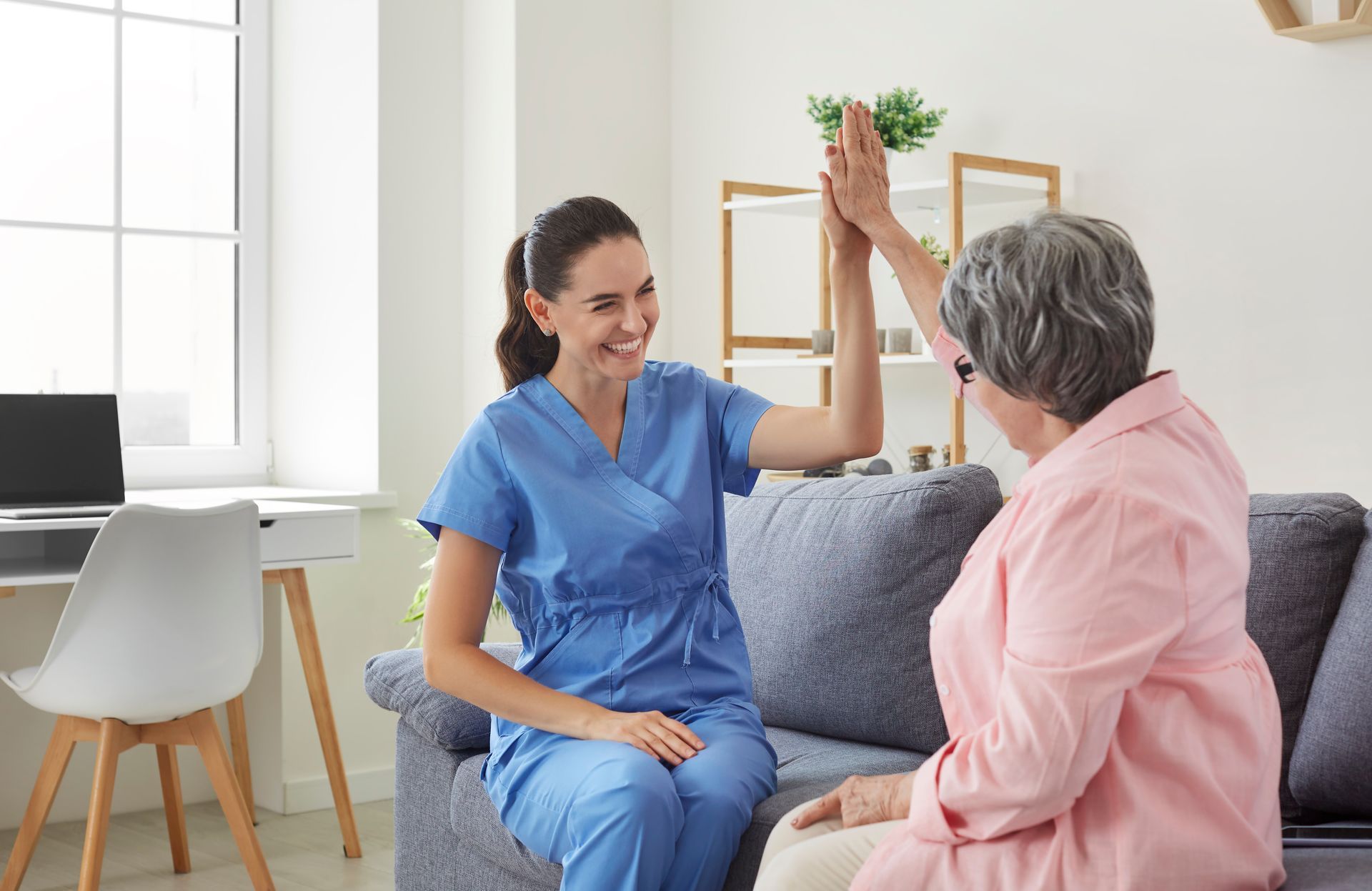 Two people high-five in a bright living room, one in blue scrubs and one seated on a gray couch.