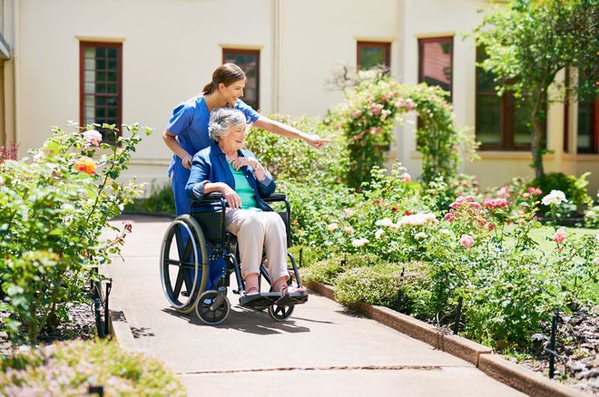 Person in a wheelchair on a garden path with a caregiver beside them, surrounded by flowers. Person in a wheelchair on a garden path with a caregiver beside them, surrounded by flowers.