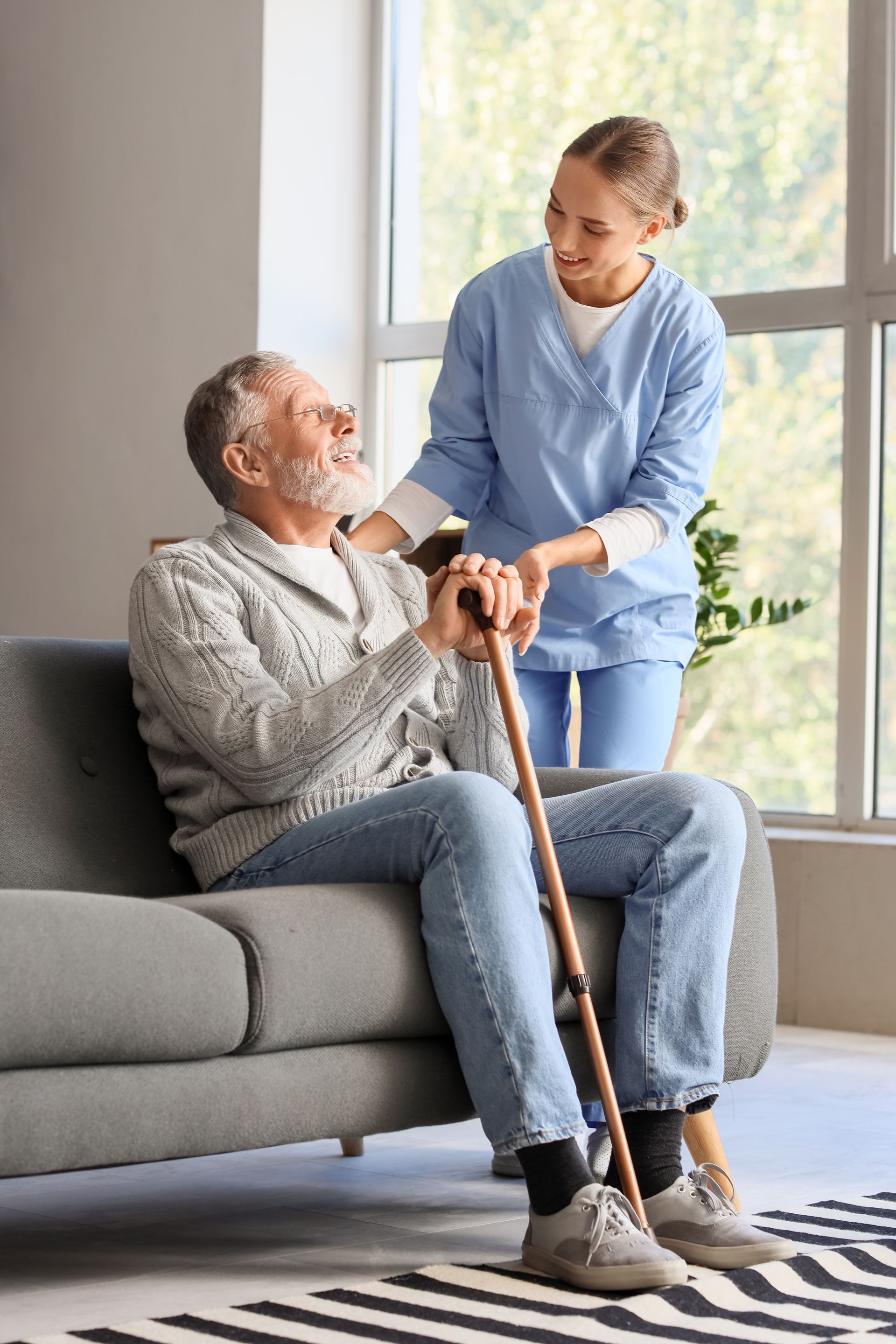 Caregiver assisting a seated man with a cane on a couch near a bright window