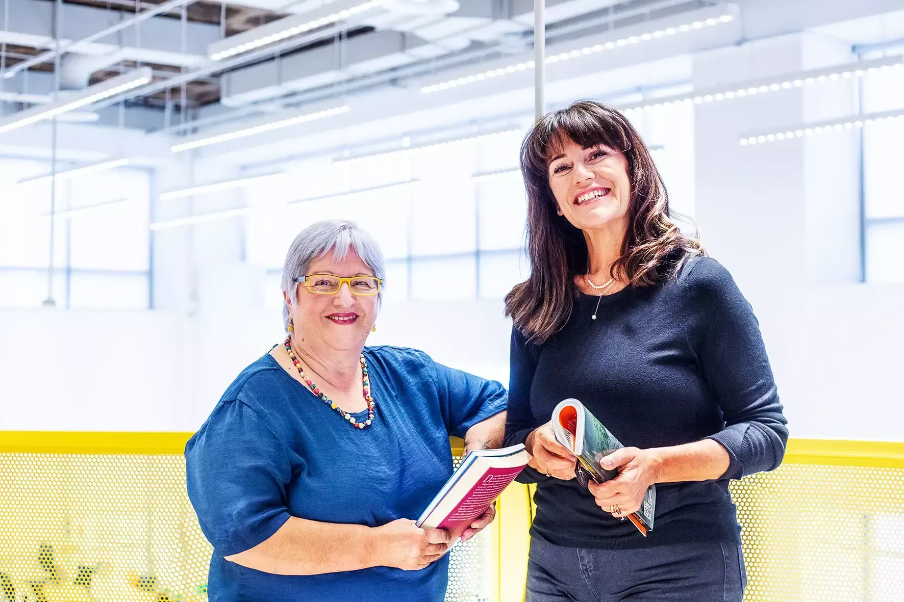Duas mulheres estão de pé, uma ao lado da outra, segurando livros.