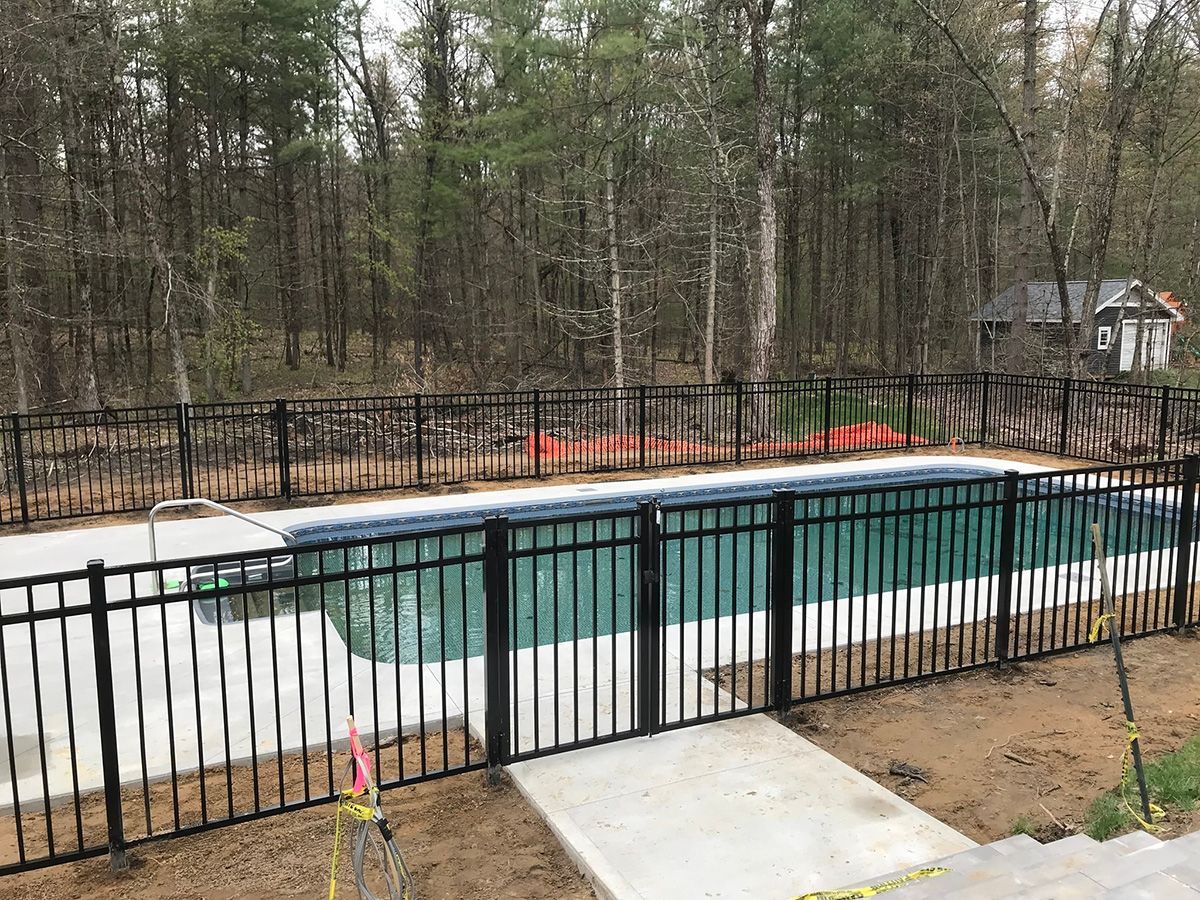 Black fenced-in rectangular pool with turquoise water. Concrete walkway. Trees in the background.
