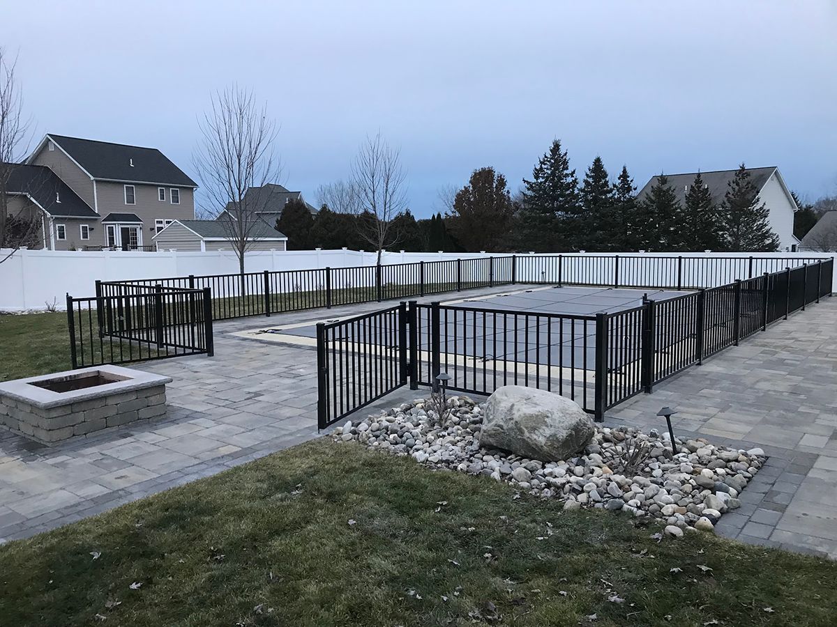 Black fenced pool area, paved patio, fire pit, large rock, and white fence. Houses in the background under cloudy sky.