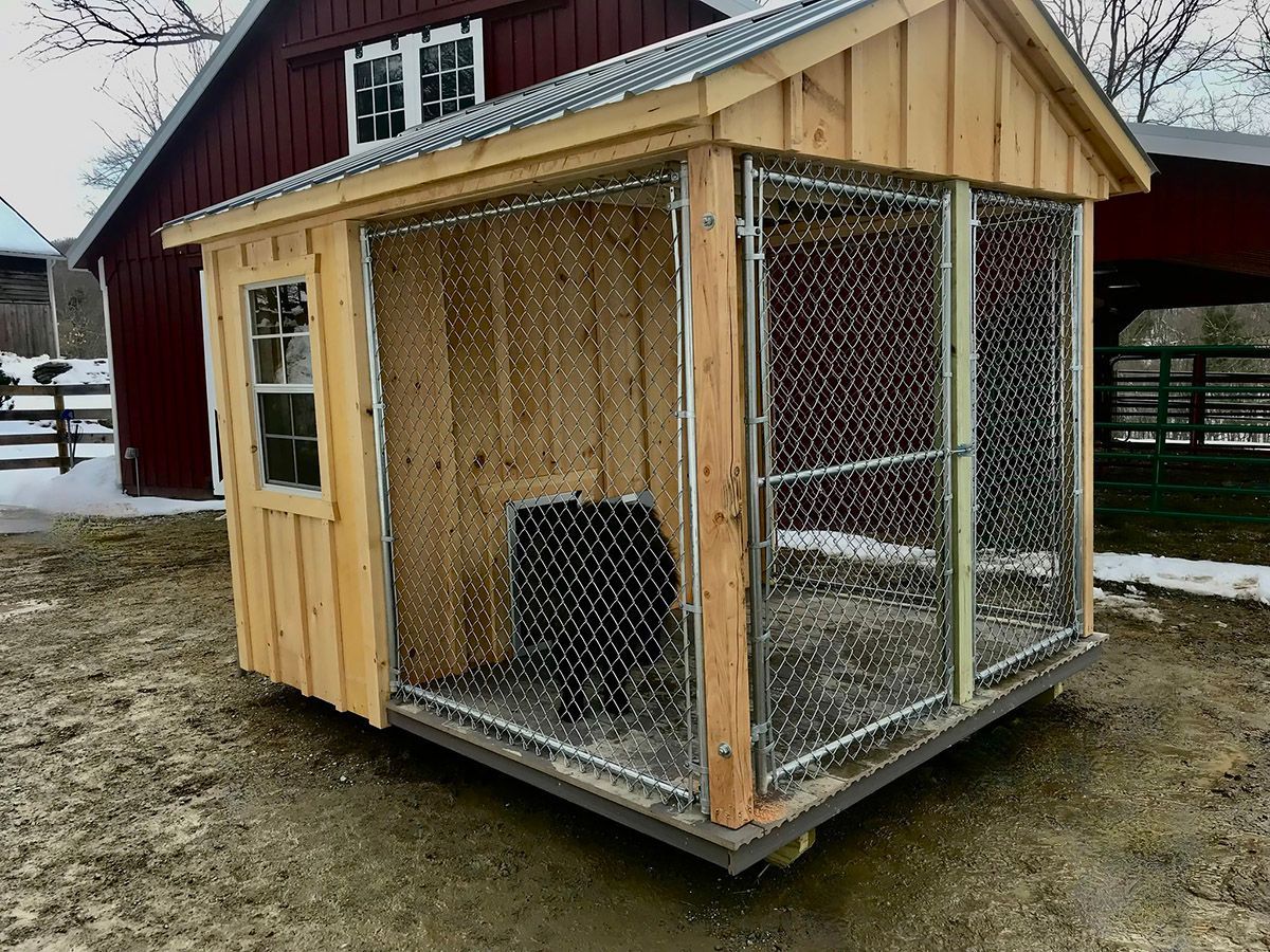 Wooden dog kennel with chain-link front, window, and metal roof, set outdoors on a muddy surface.