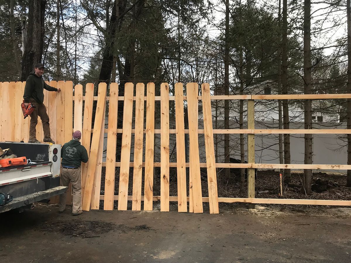 Two workers building a wooden fence outdoors, one on a truck, other on the ground.