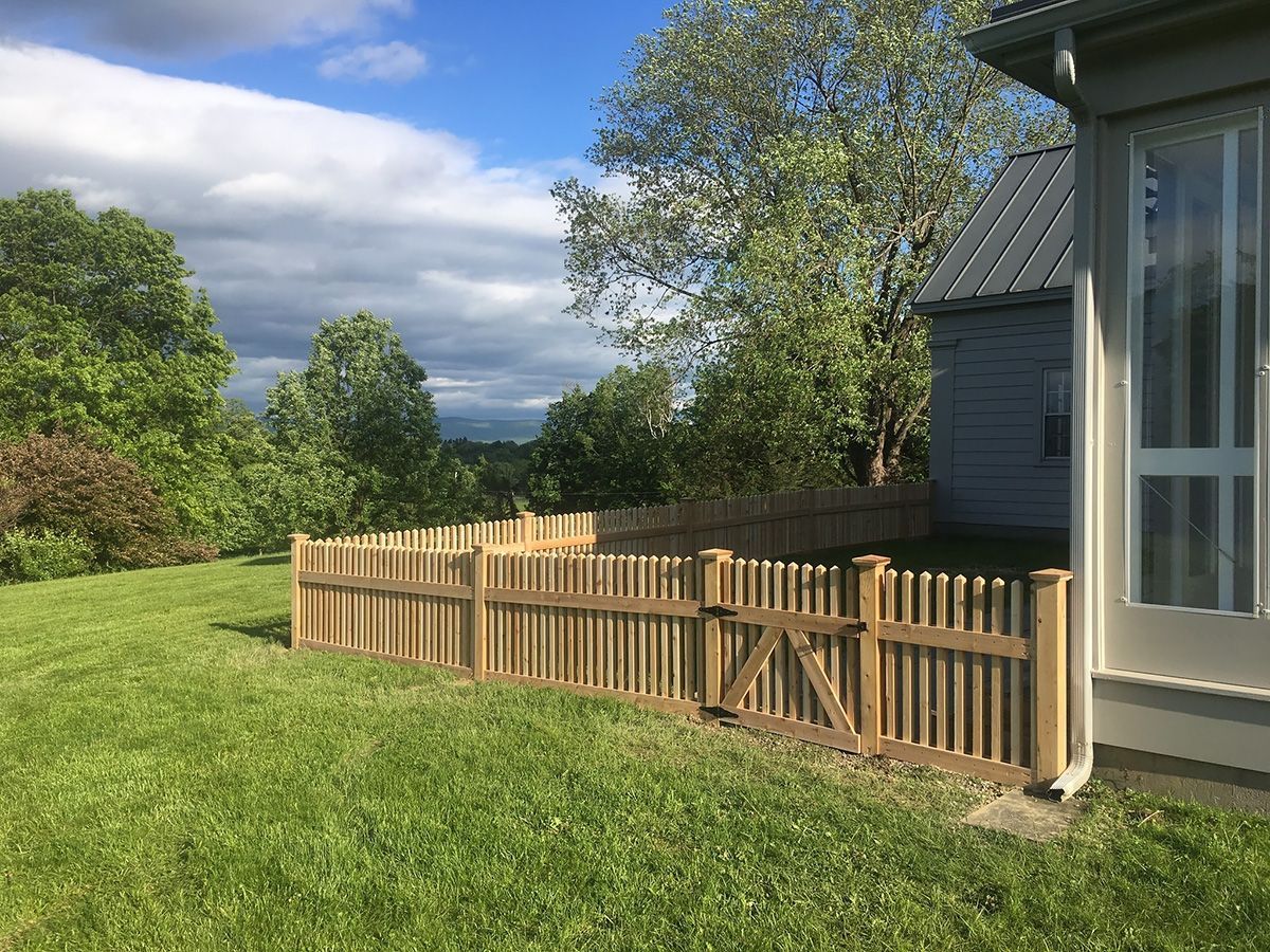 Wooden picket fence in a grassy yard, beside a house with a window, under a sunny sky.