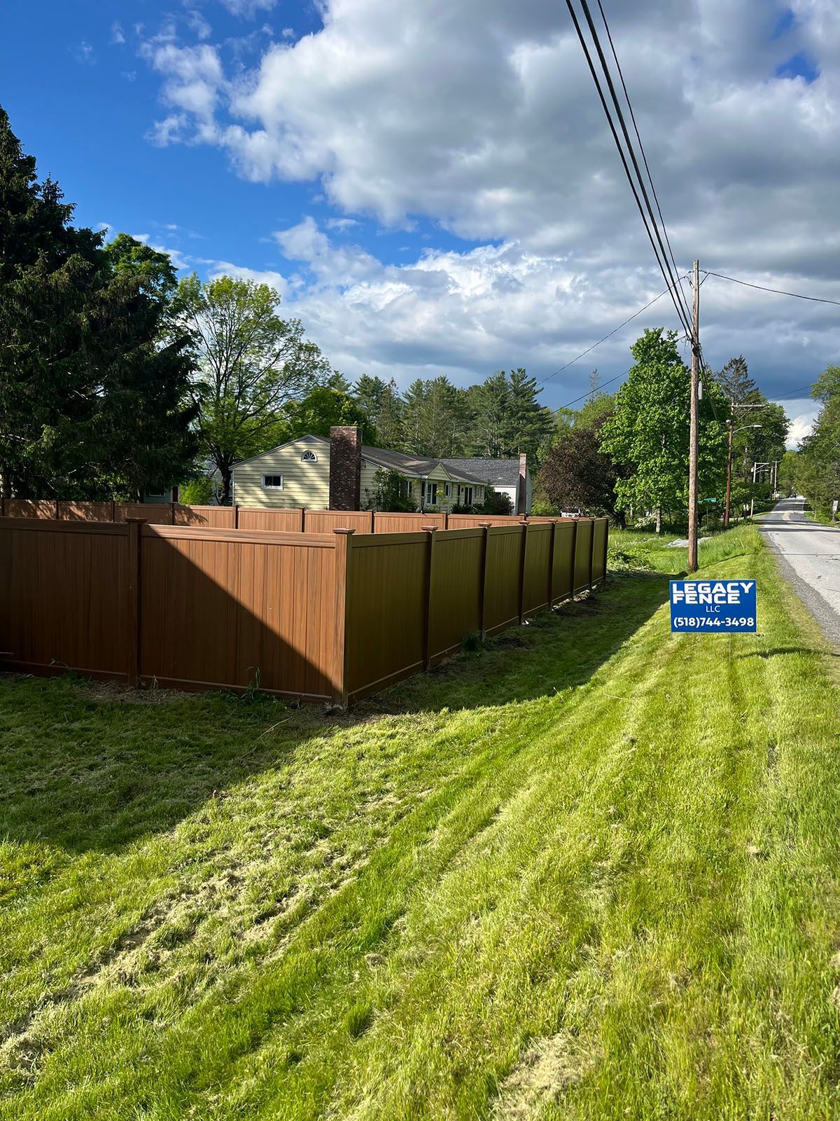 Brown privacy fence bordering a grassy lawn, with a house visible in the background under a blue sky.
