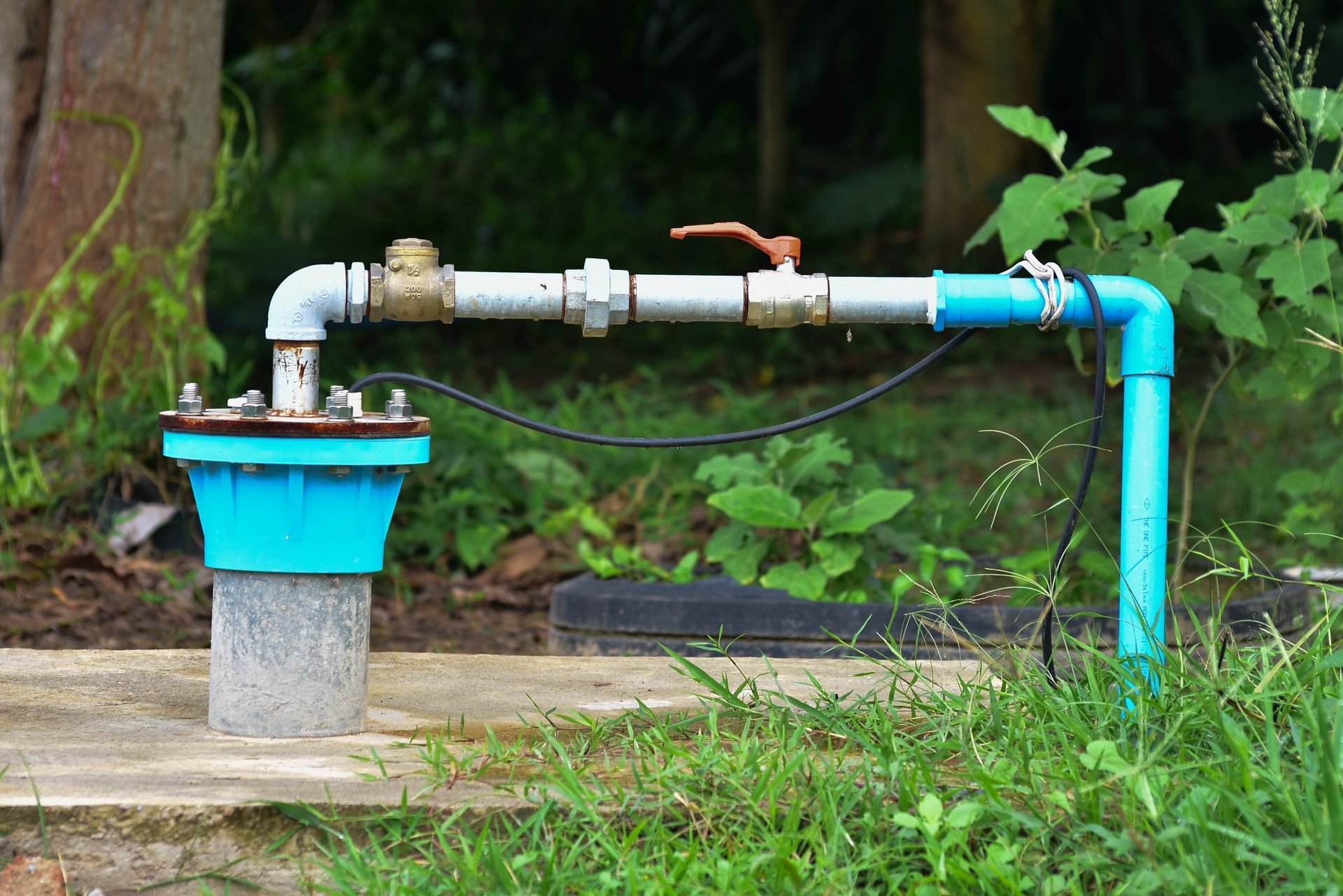 Outdoor wellhead with blue PVC piping, metal valve, and pump connection. Outdoor wellhead with blue PVC piping, metal valve, and pump connection.