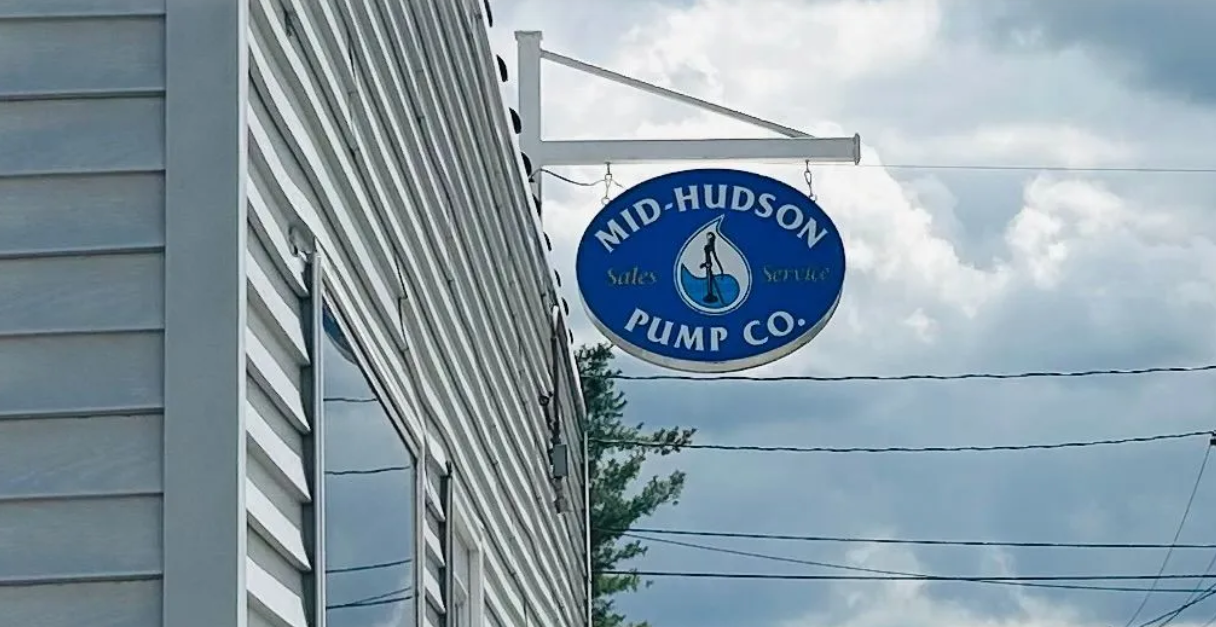 Blue circular sign for Mid-Hudson Pump Co. hangs from a white bracket on a building's side.