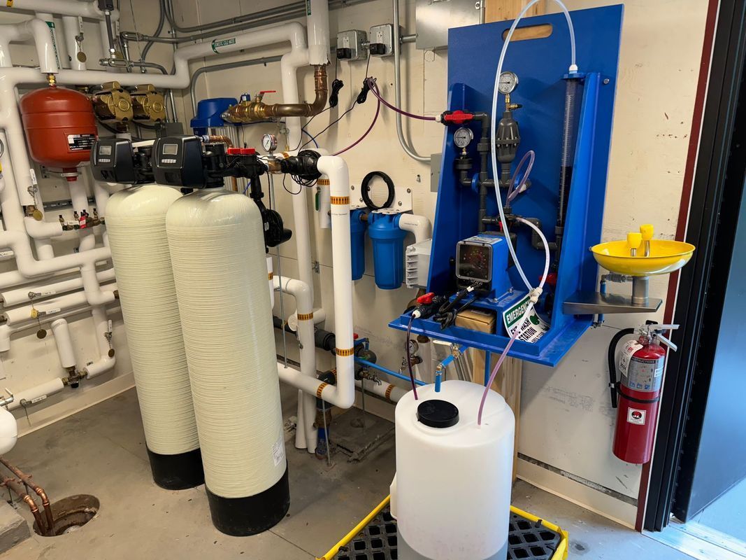 Water filtration and control system with tanks, pipes, and a blue panel with electrical components in a mechanical room.