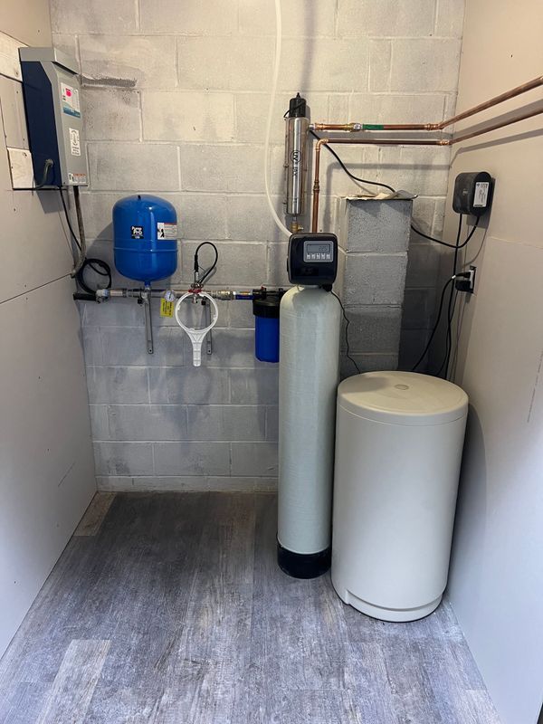 Water treatment system in a utility room; features a tank, filter, pressure tank, and pipes against a concrete block wall.