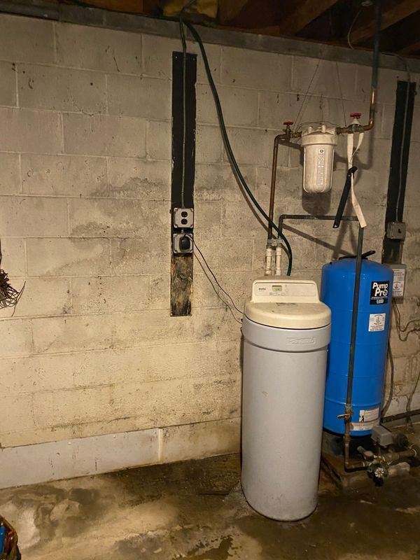 Water softener and filter system in a basement setting. Gray and blue tanks, white filter housing, on a concrete block wall.