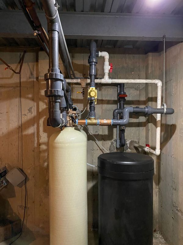 Water filtration system with two tanks and connected pipes against a concrete wall.