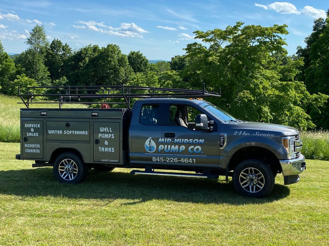 Service truck parked in a grassy field. Truck is gray, with company name, phone number, and services listed on the side. Blue sky and trees in the background.