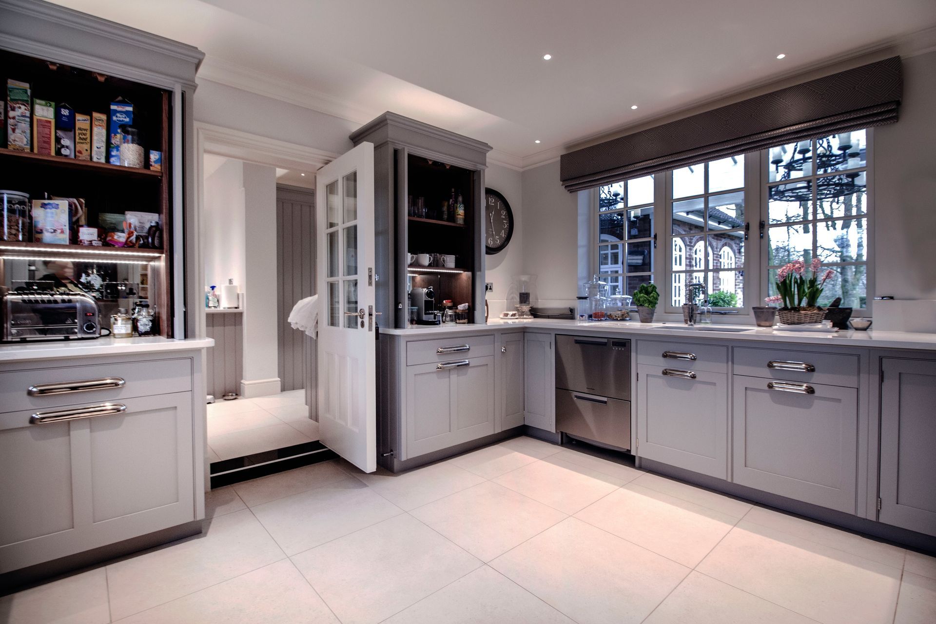 A modern kitchen with grey cabinets, light tile flooring, stainless steel appliances, and a view through a doorway.
