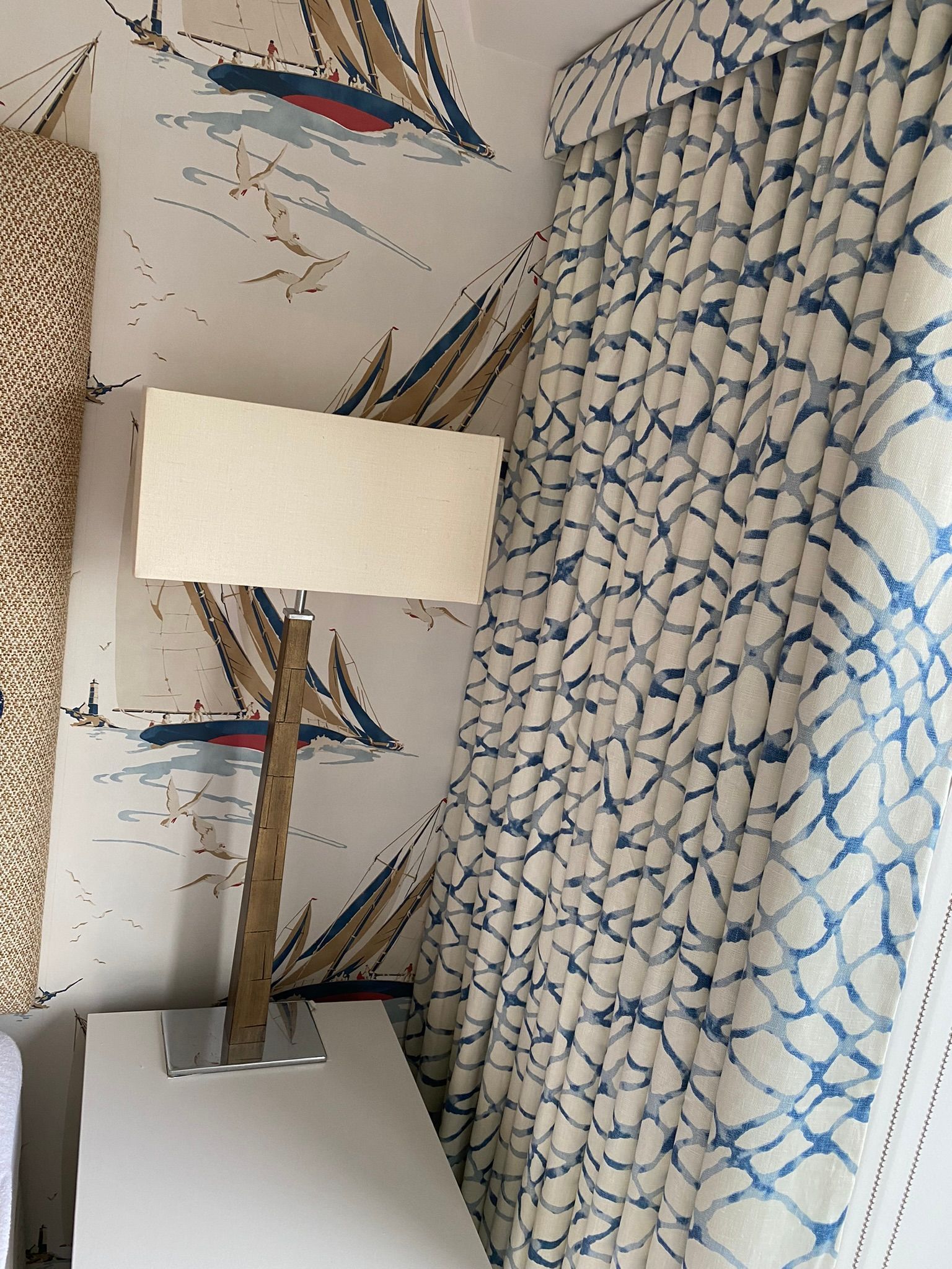 A tall lamp with a textured stand sits on a white surface next to blue patterned curtains and sailboat-themed wallpaper.