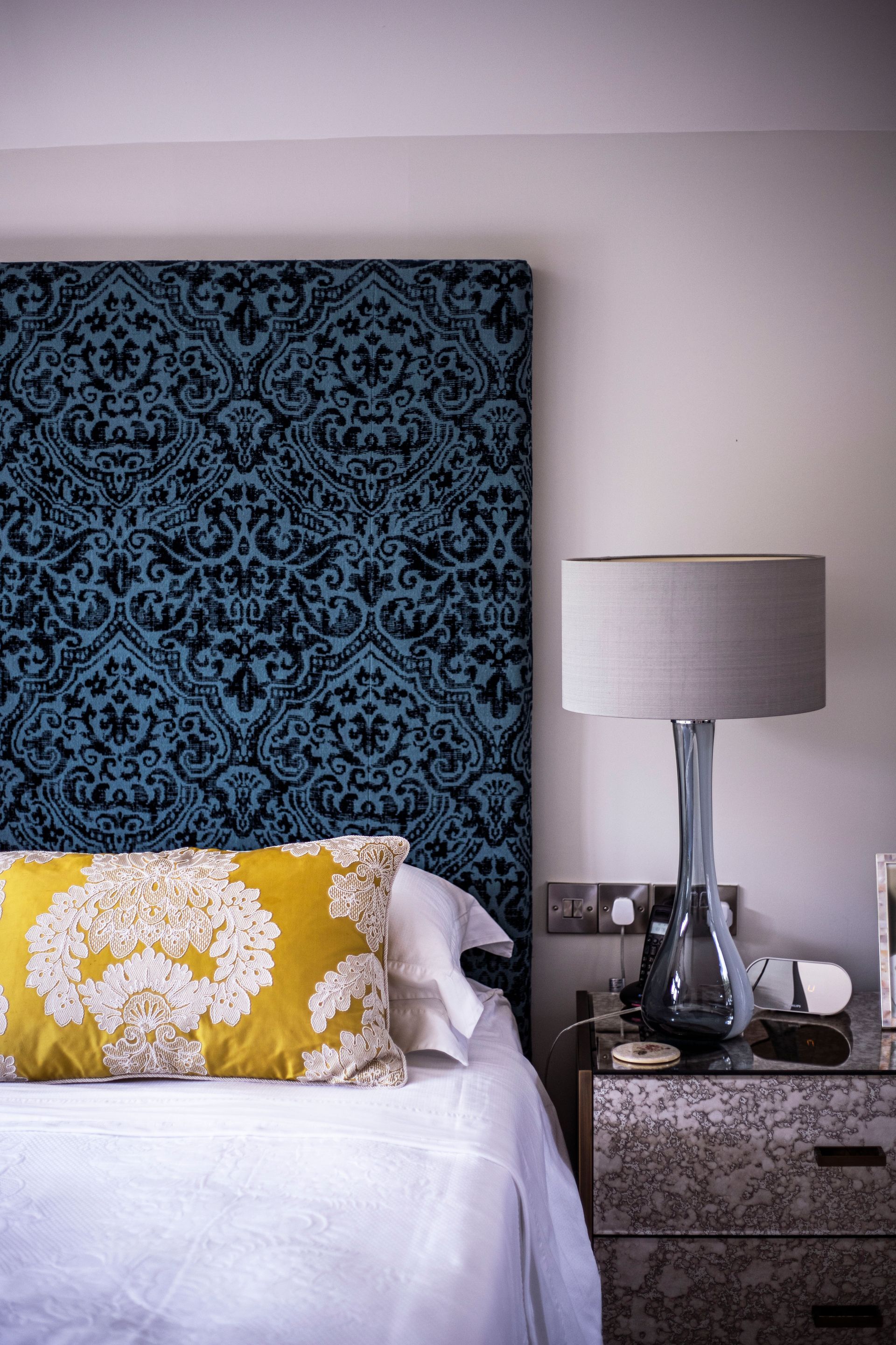 A bedroom corner featuring a blue-patterned headboard, a yellow floral accent pillow, and a lamp on a patterned nightstand.