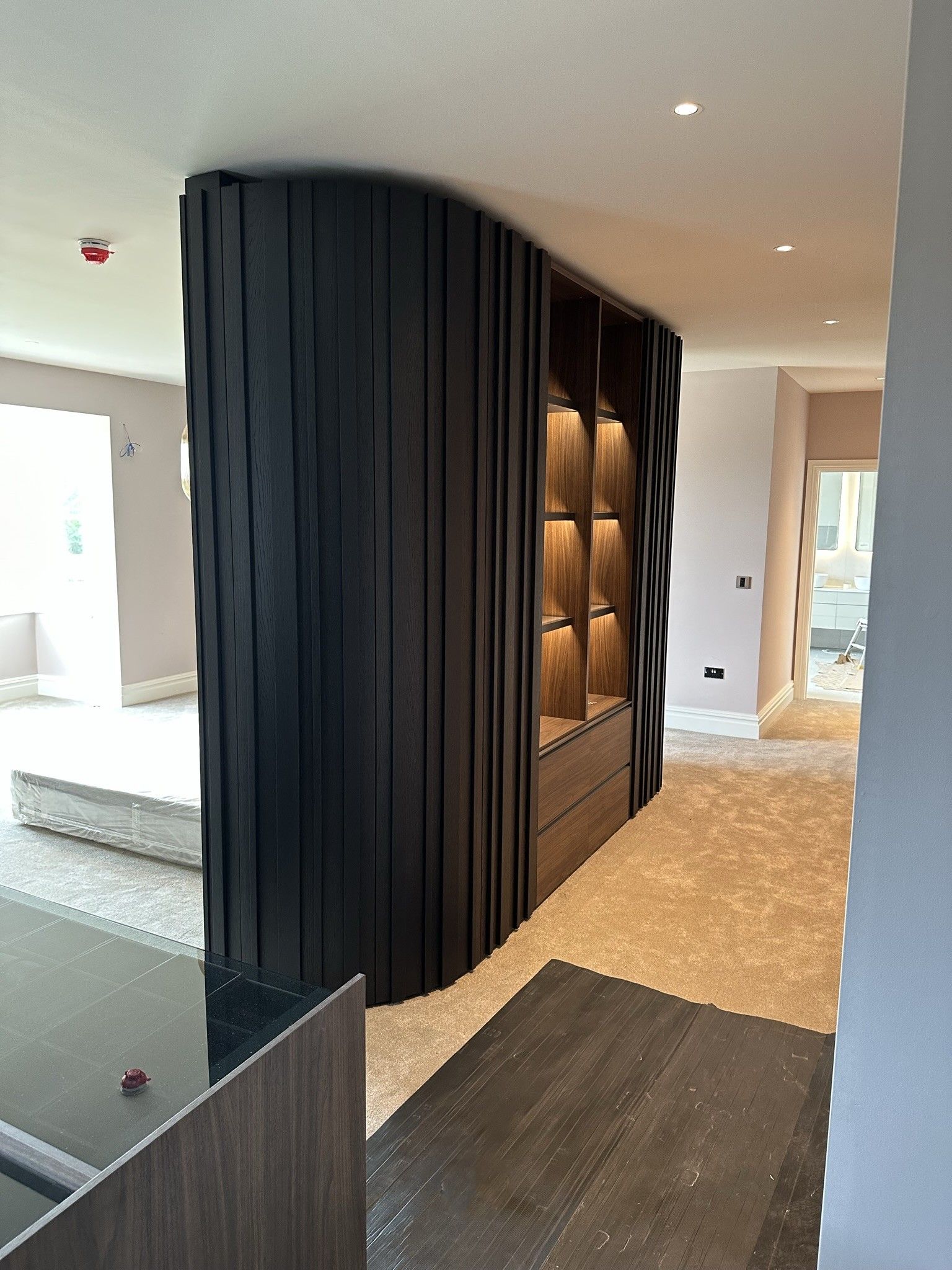 A curved, black vertical-slatted room divider with built-in, illuminated wooden shelving in a modern interior.