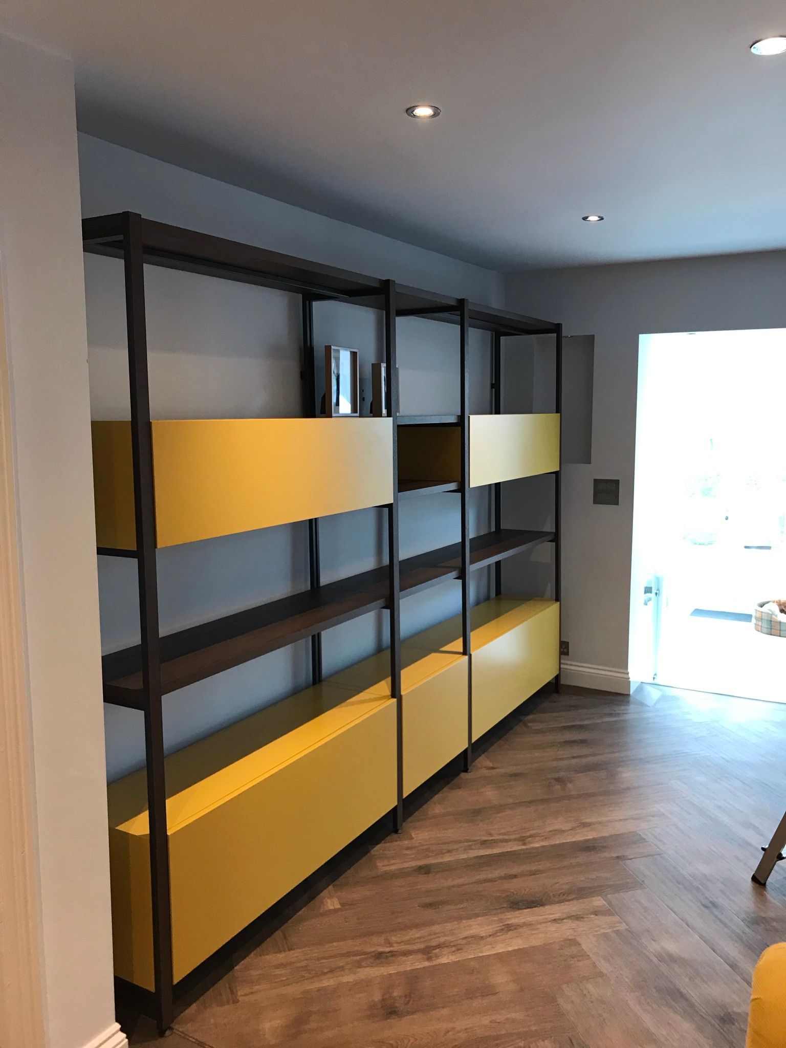 Tall, industrial-style black metal shelving unit with yellow storage cabinets on a herringbone wood floor in a room.