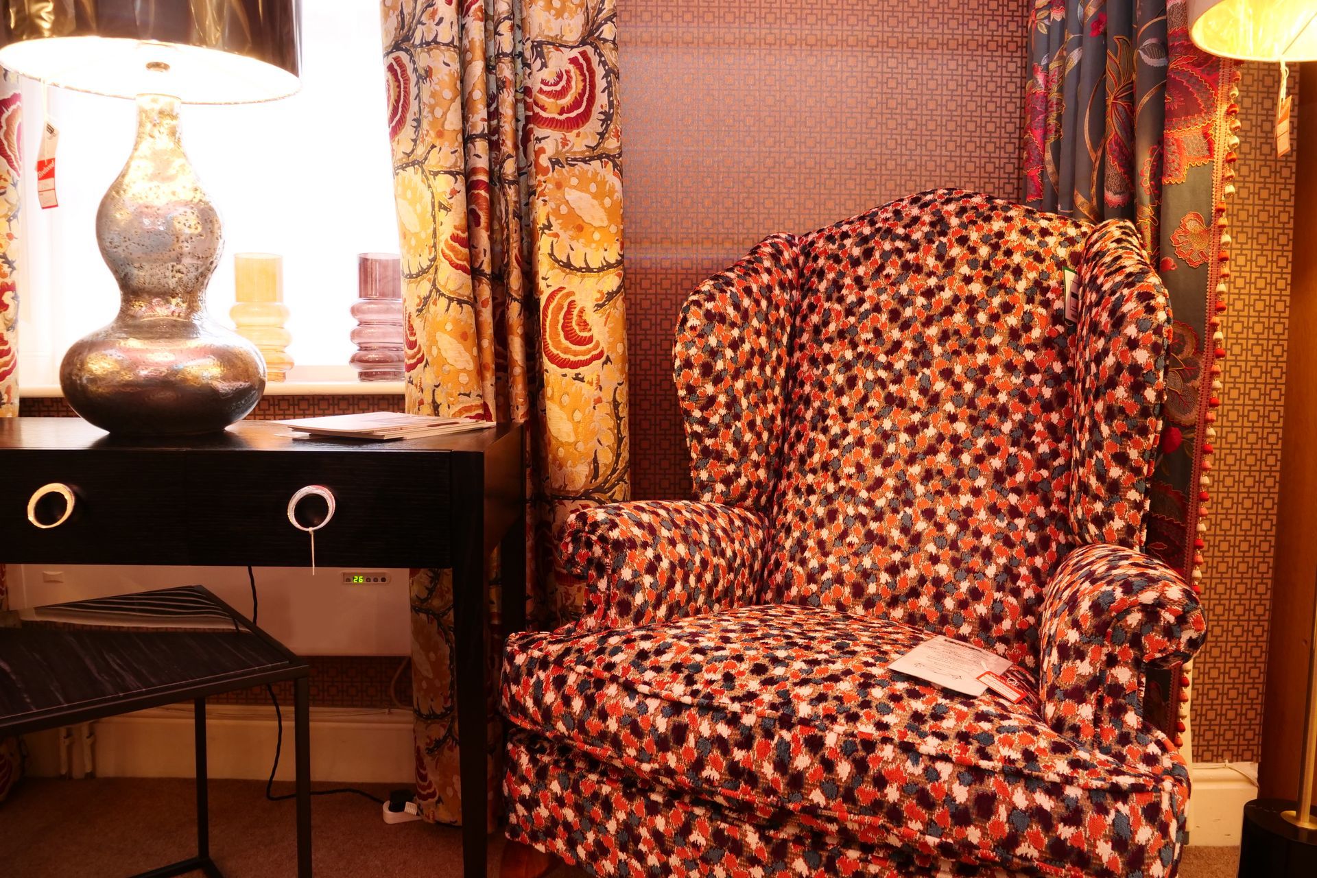 A wingback chair with a colorful, patterned fabric sits next to a black desk with a lamp in a cozy, decorated room.