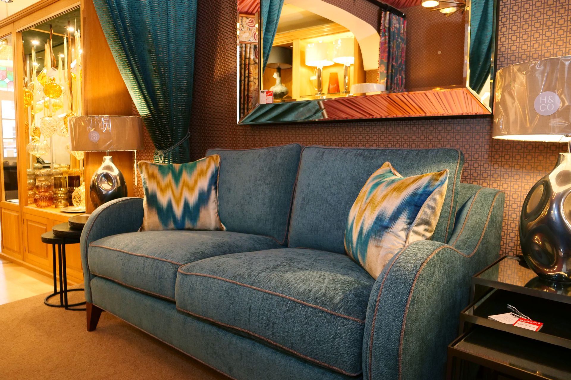 A teal fabric sofa with patterned accent pillows sits in a warmly lit room with a mirror and lamps.