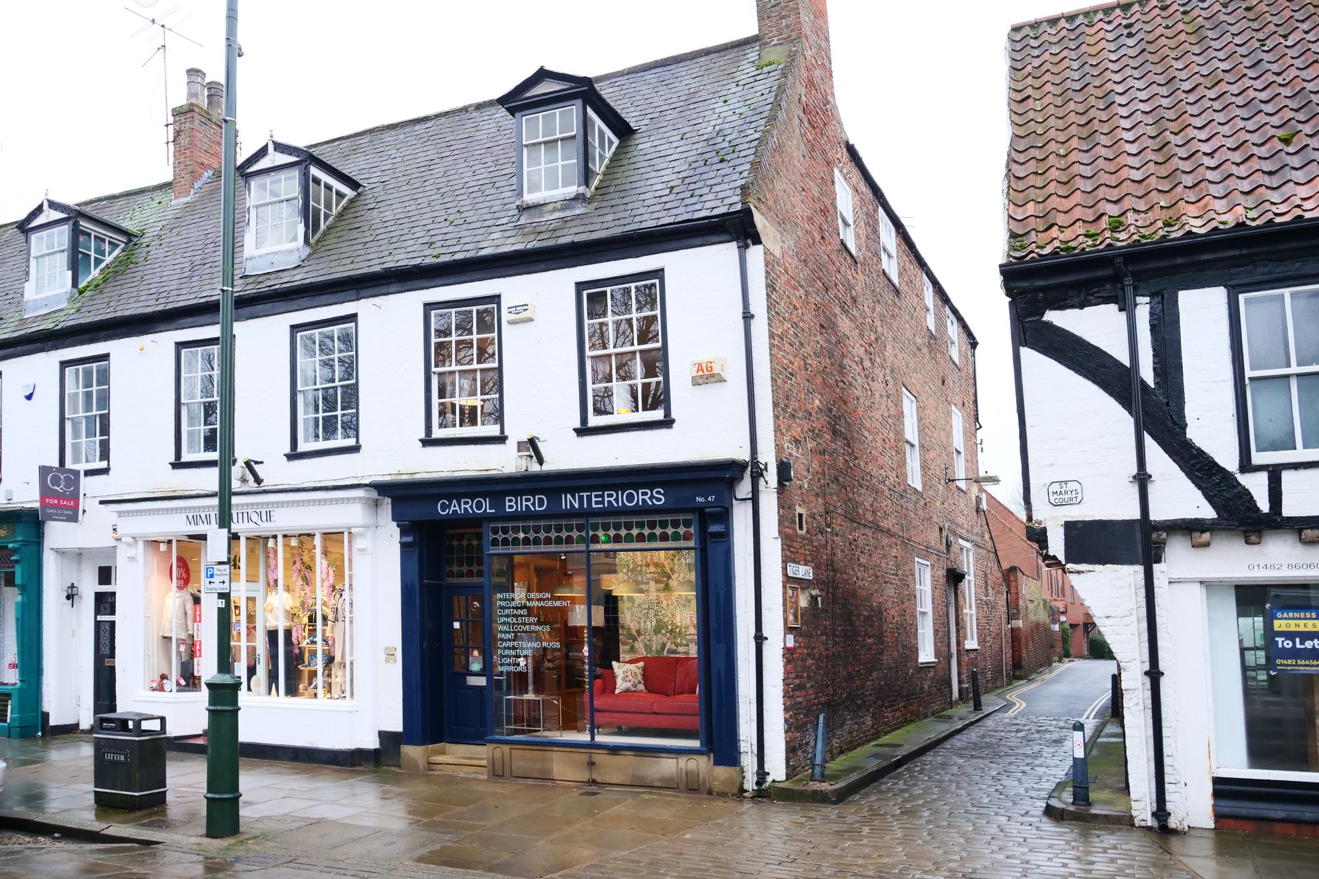 A street-level view of a white, multi-story building featuring shops on the ground floor next to a cobblestone alleyway.
