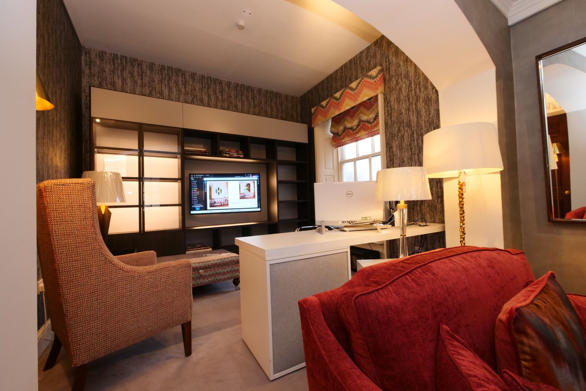 A room with a red sofa, a patterned armchair, a built-in media unit with a television, and a white desk under a window.