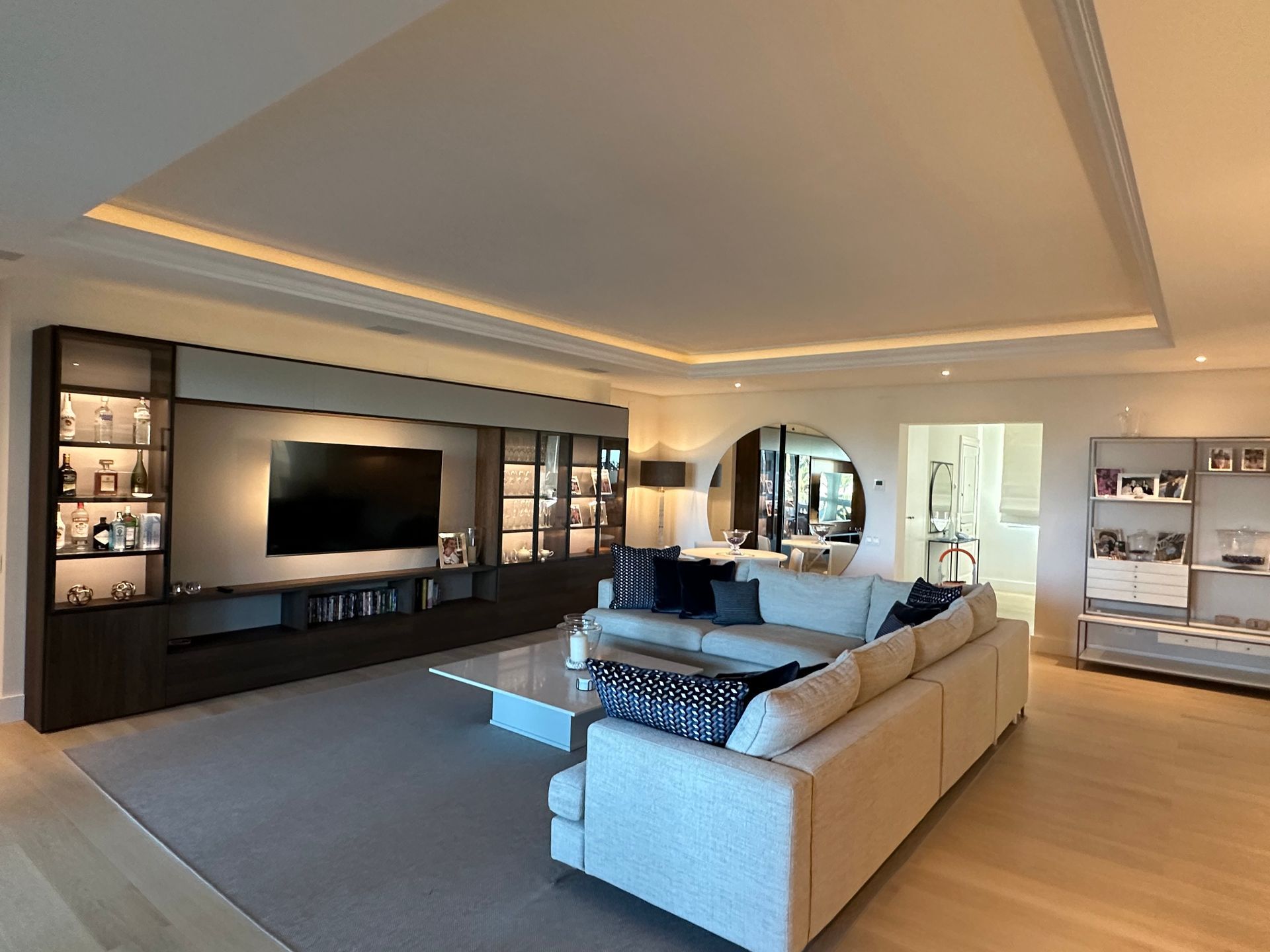 A modern living room with a built-in media wall, a large cream sectional sofa on a gray rug, and light wood flooring.