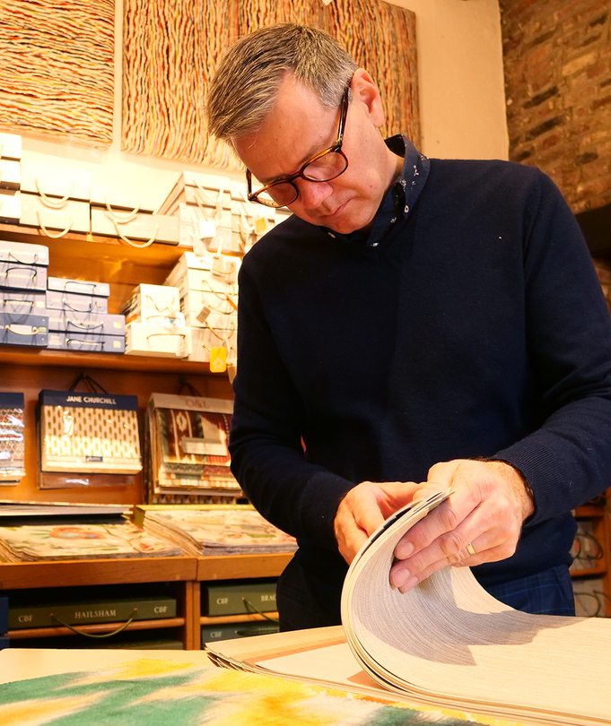 A person in a dark sweater examines fabric samples at a table in a shop filled with wallpaper or textile books.