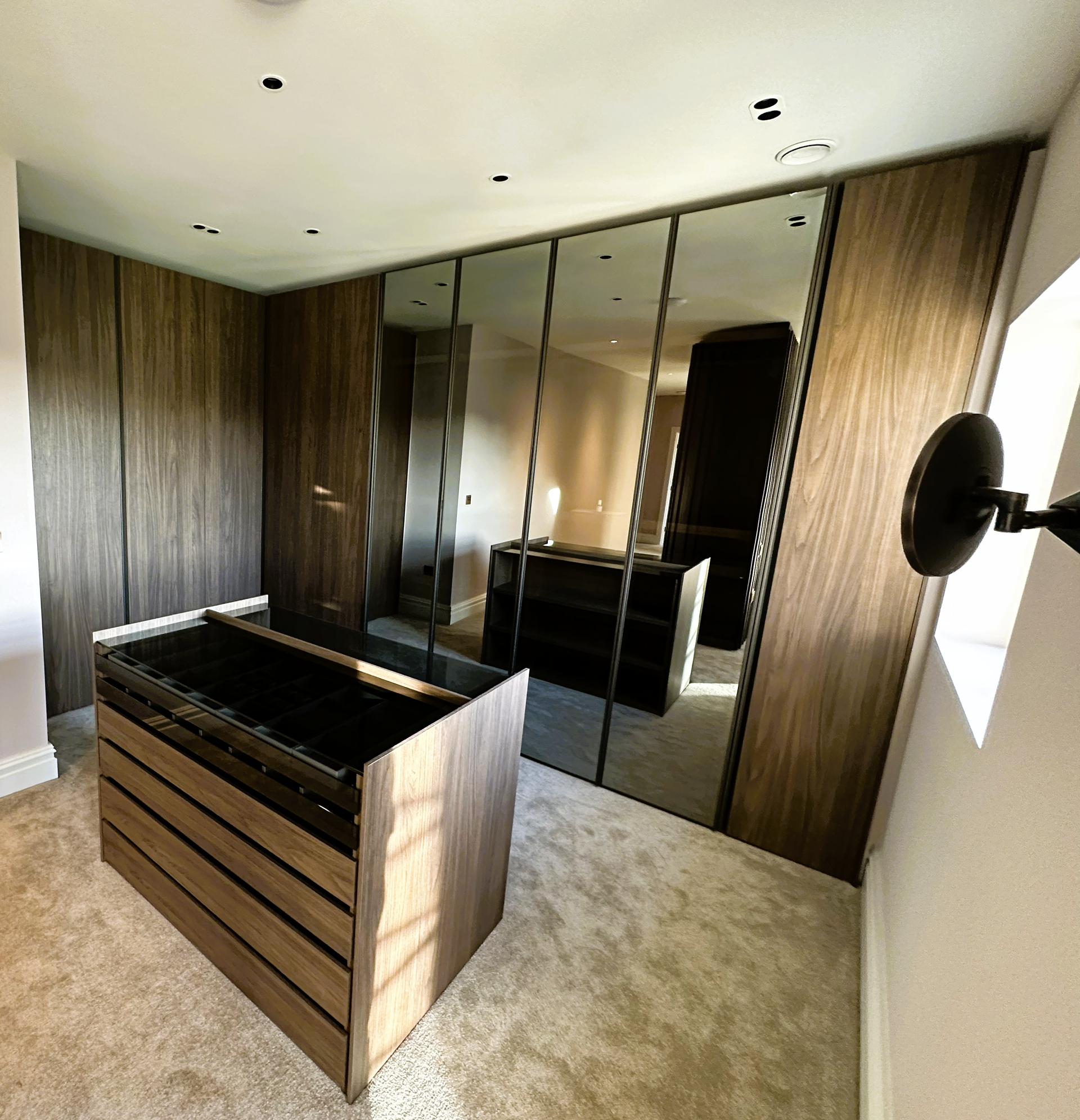 A walk-in closet with dark wood-paneled cabinets, mirrored wardrobe doors, and a matching central storage island unit.