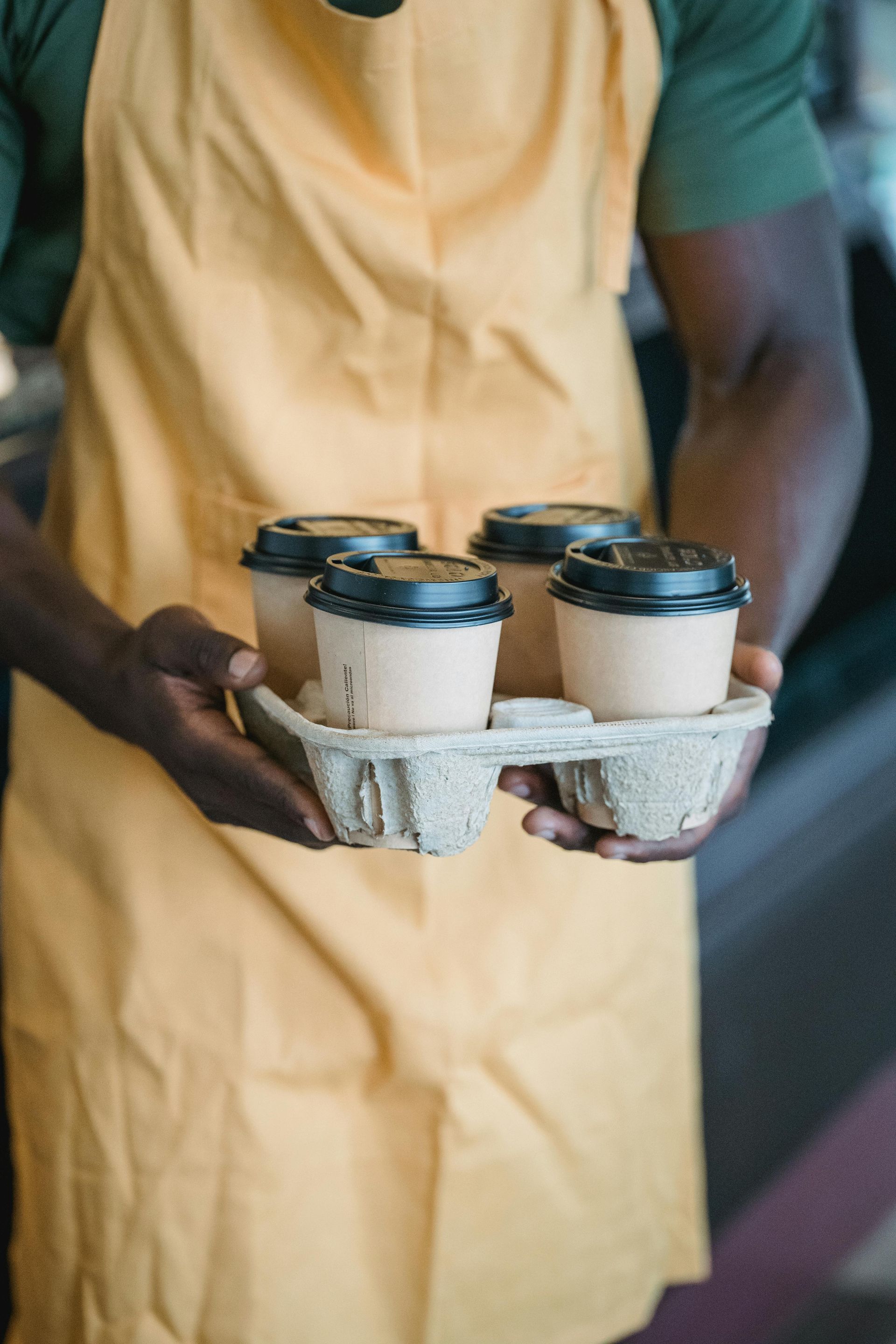 Person in yellow apron holding a tray with four coffee cups.