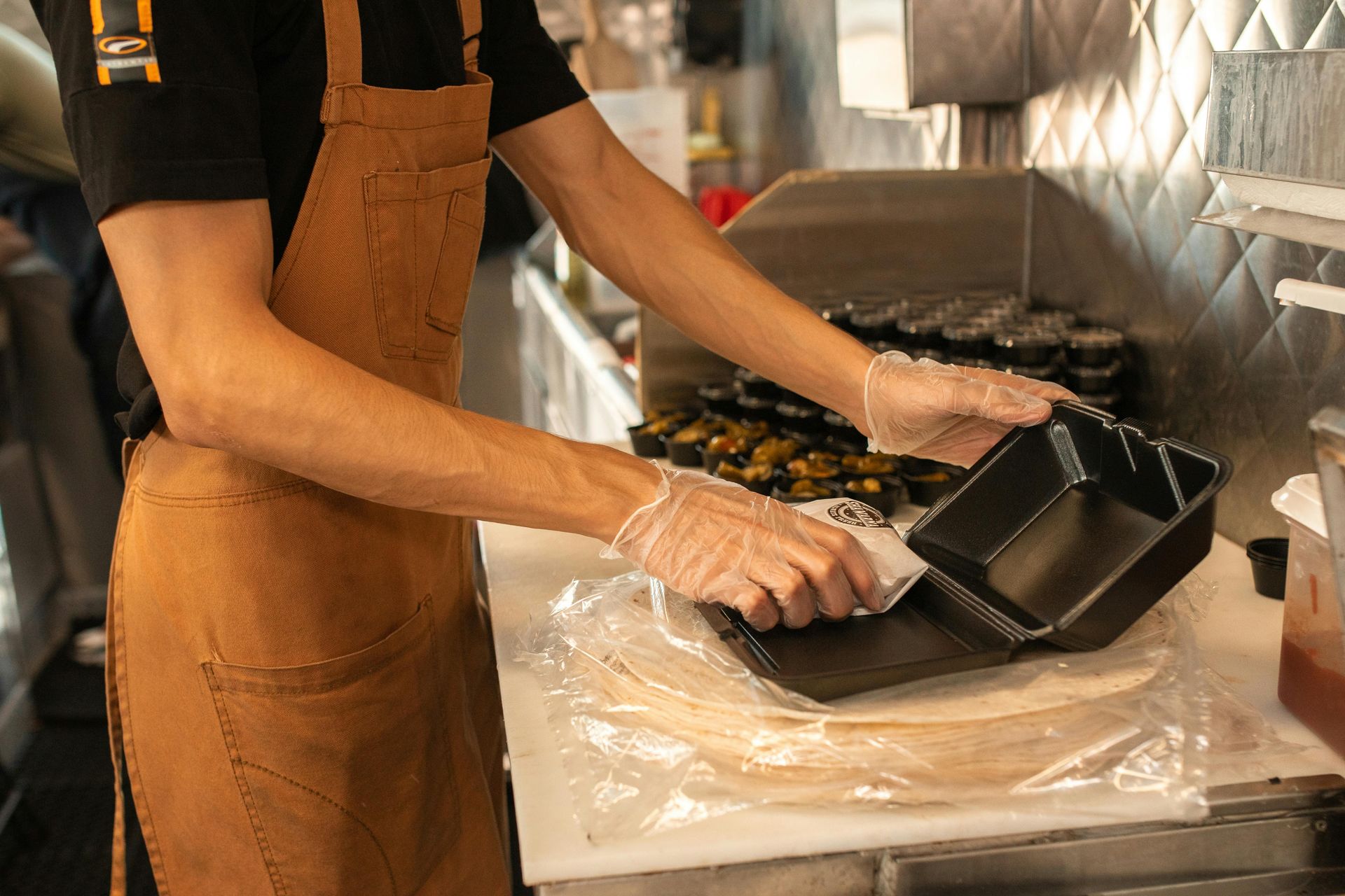 Person in apron prepares food in a food truck, placing items into a black container.