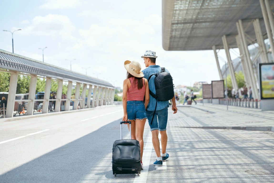 Dos viajeros con mochilas caminan por un aeropuerto moderno, arrastrando una maleta negra.