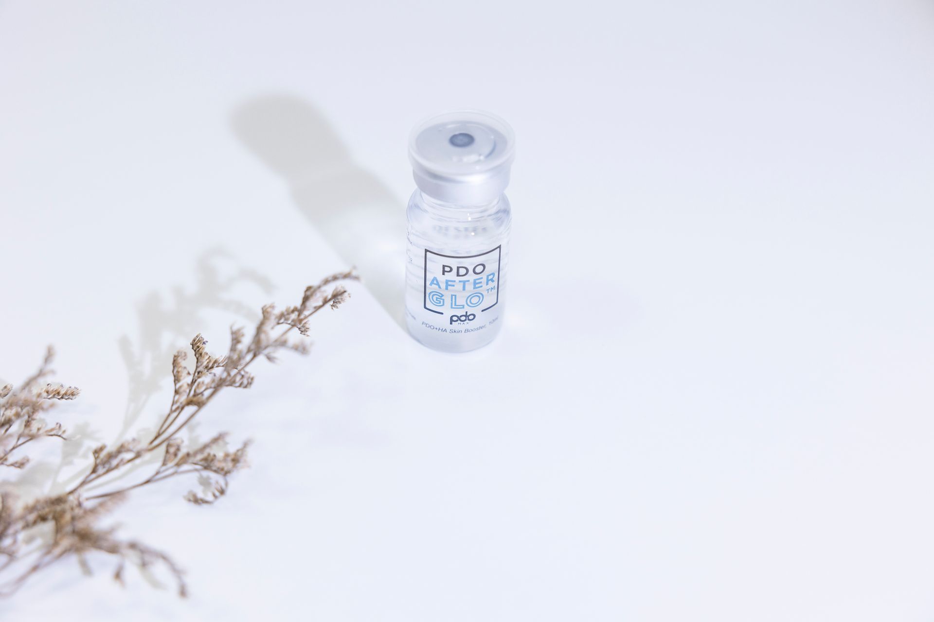 A small, clear vial with a label, on a white surface with a dried flower.