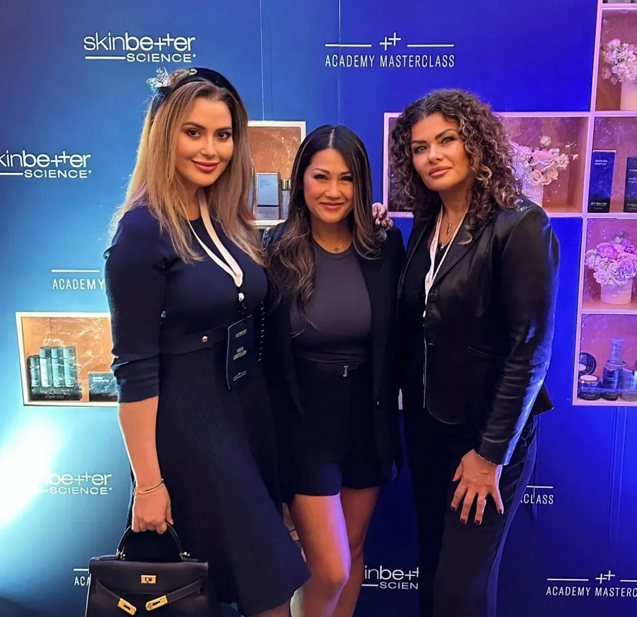 Three women pose in front of a blue backdrop, some wearing name tags. One holds a black handbag.