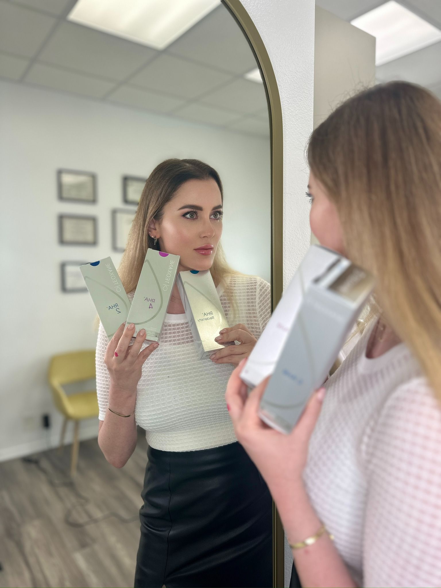 Woman holding product boxes in front of a mirror, looking at reflection. White room, gold mirror frame.