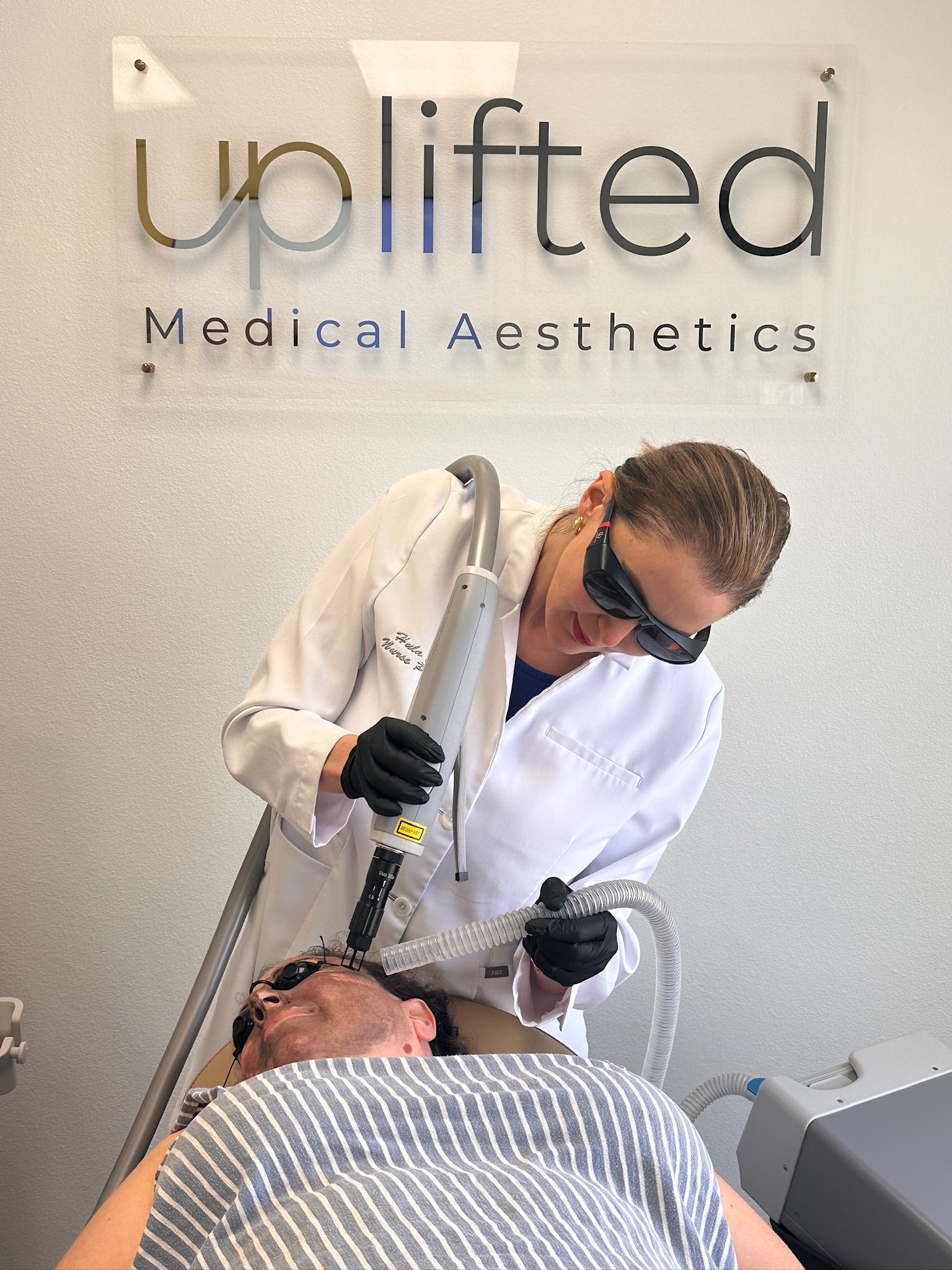 Hula Castellon, FNP, MSN, and owner of Uplifted Aesthetics using a medical device on a patient's face.