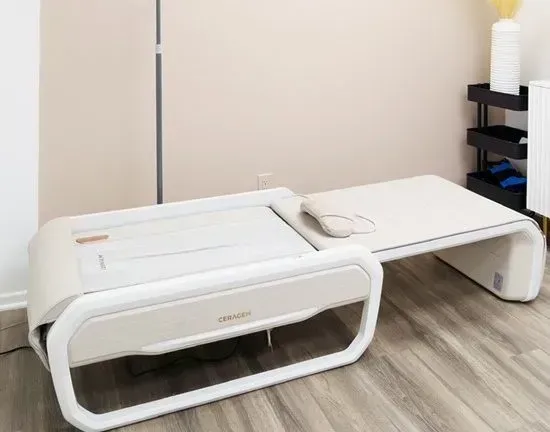 Beige massage bed in a light-colored room. Includes a pillow and small side table with items.