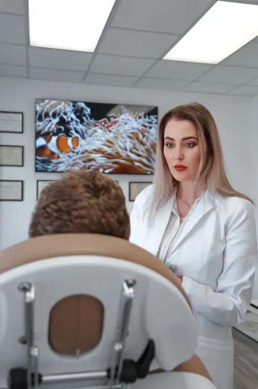 Hula Castellon, FNP, MSN, and owner of Uplifted Aesthetics speaking to a patient in an examination chair; clinic interior.