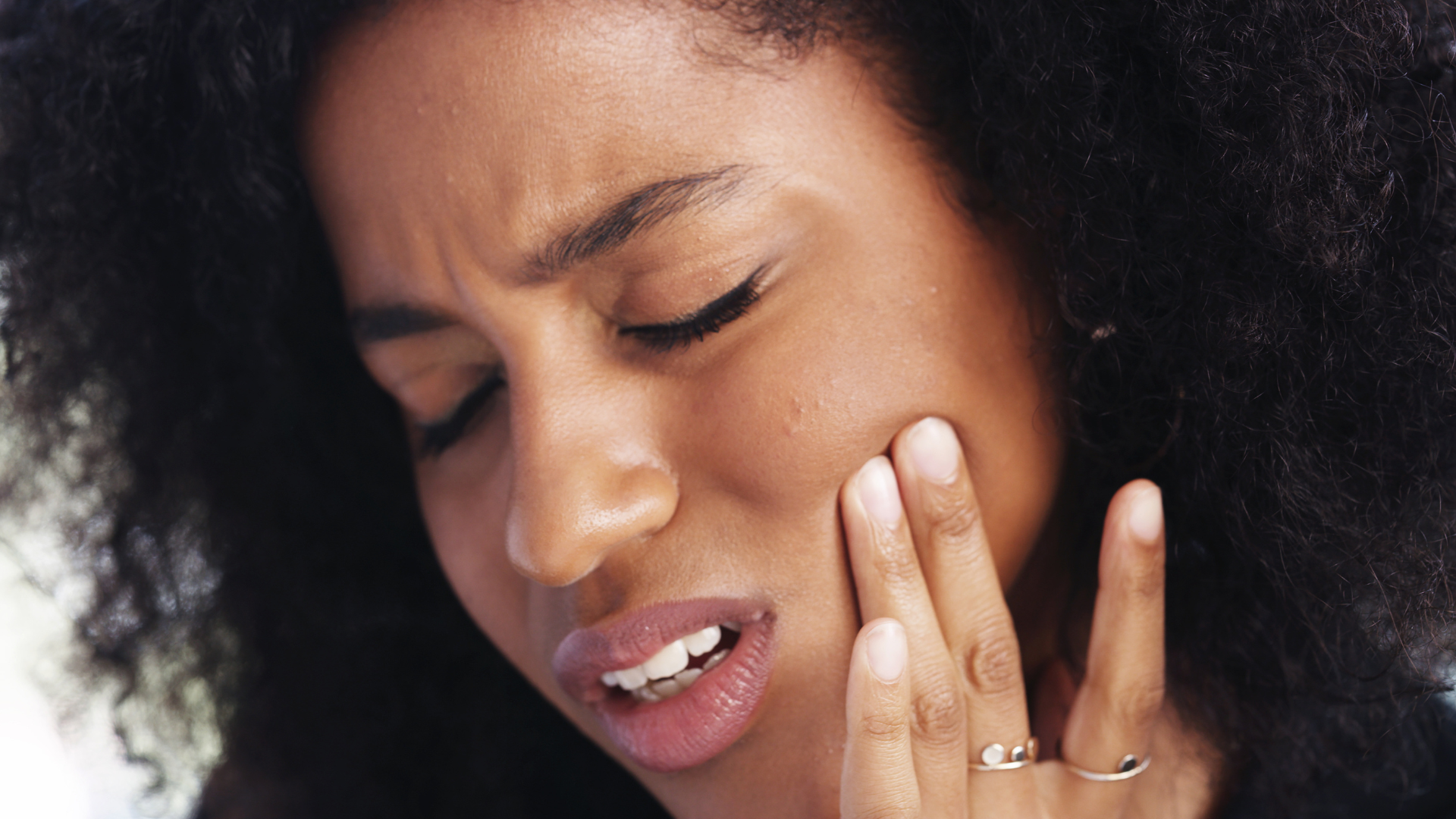 Woman with curly hair grimacing and touching cheek, experiencing TMJ pain before her treatment.