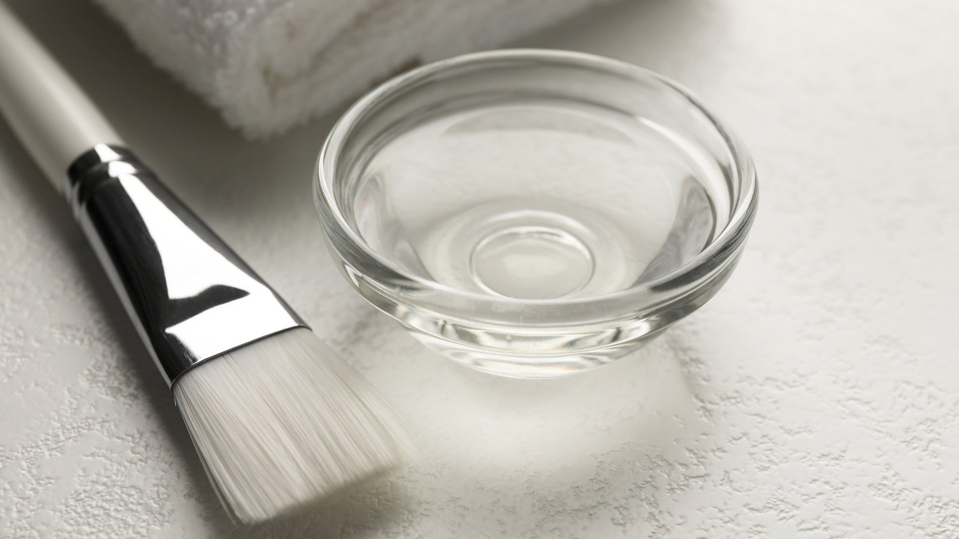 A makeup brush, small glass bowl with liquid, and a folded white towel on a light surface.