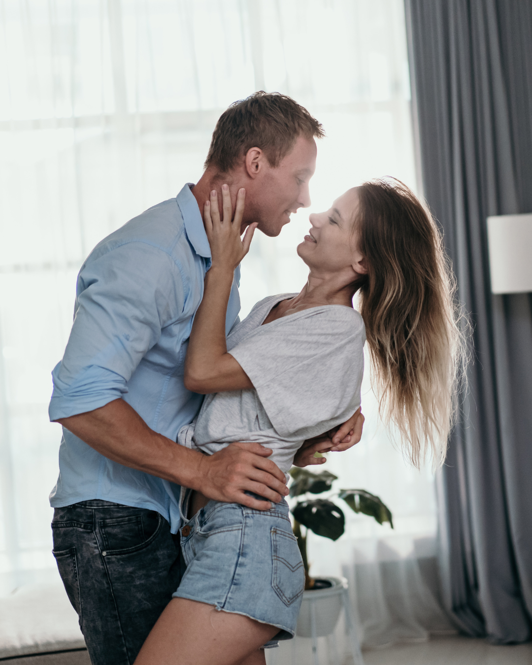 Couple embracing, smiling, looking at each other. Man in blue shirt, jeans; woman in t-shirt, shorts. Inside, by window.