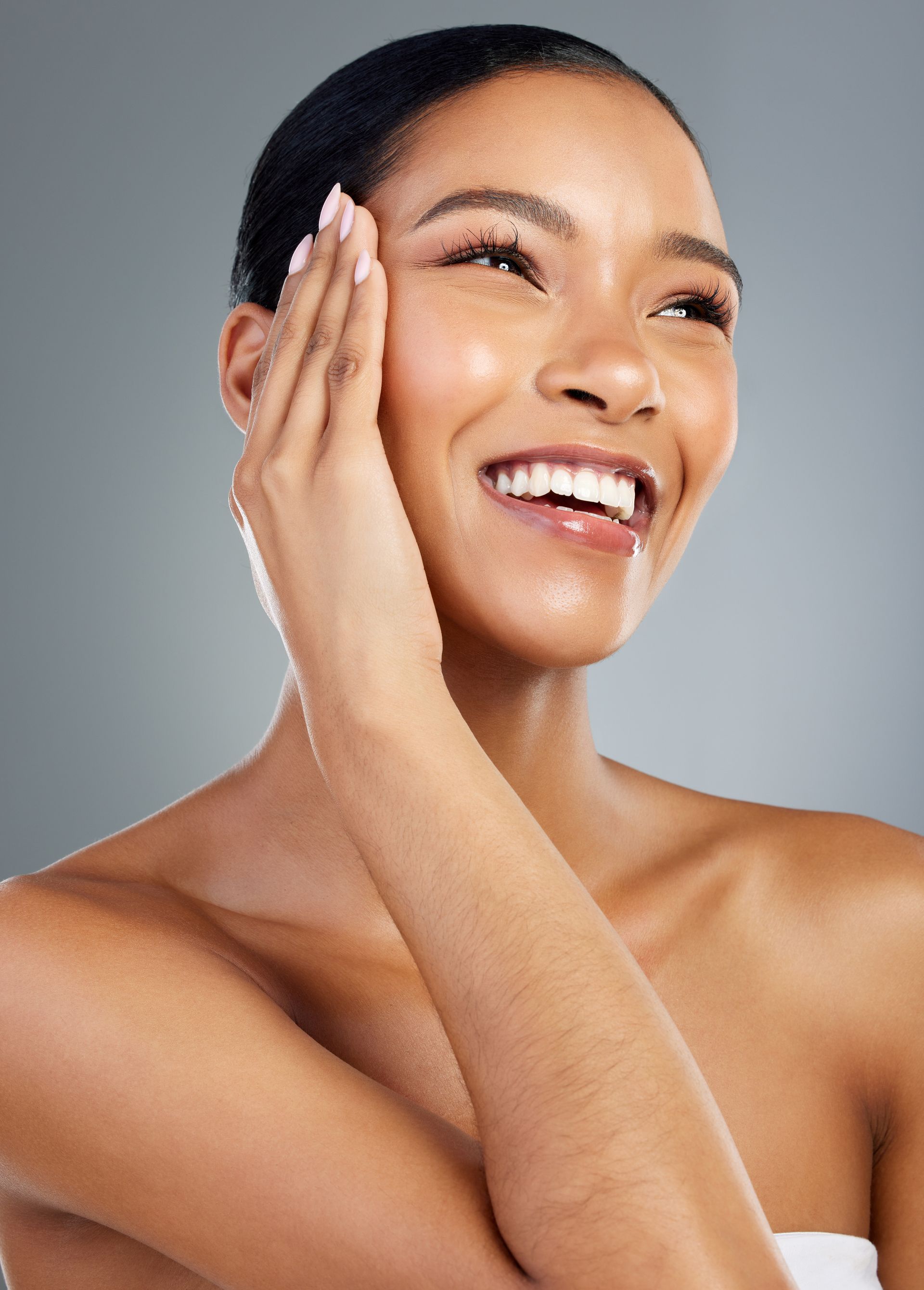 Woman smiling, touching face, illuminated skin, against a gray background.