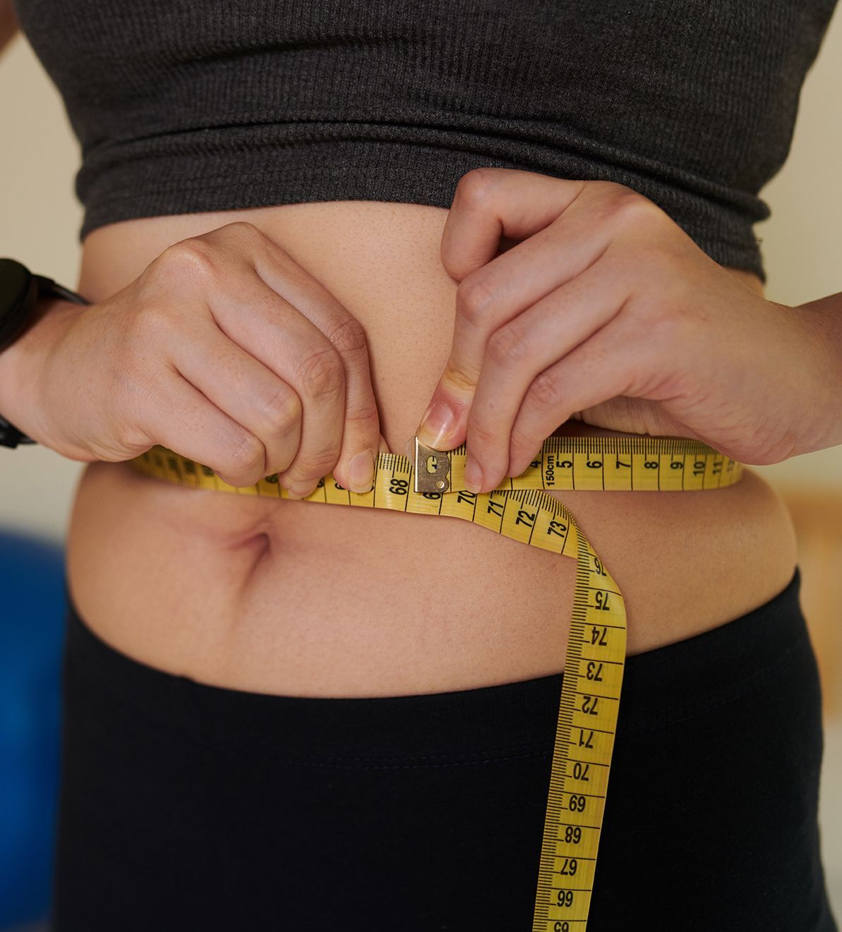 Person measuring waist with a yellow tape measure, stomach visible, wearing a gray top and black pants.