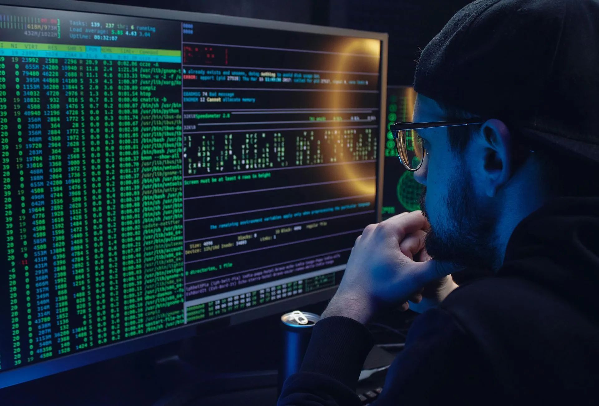 A person wearing a cap and glasses looks intently at a computer screen filled with lines of glowing code in a dark room.
