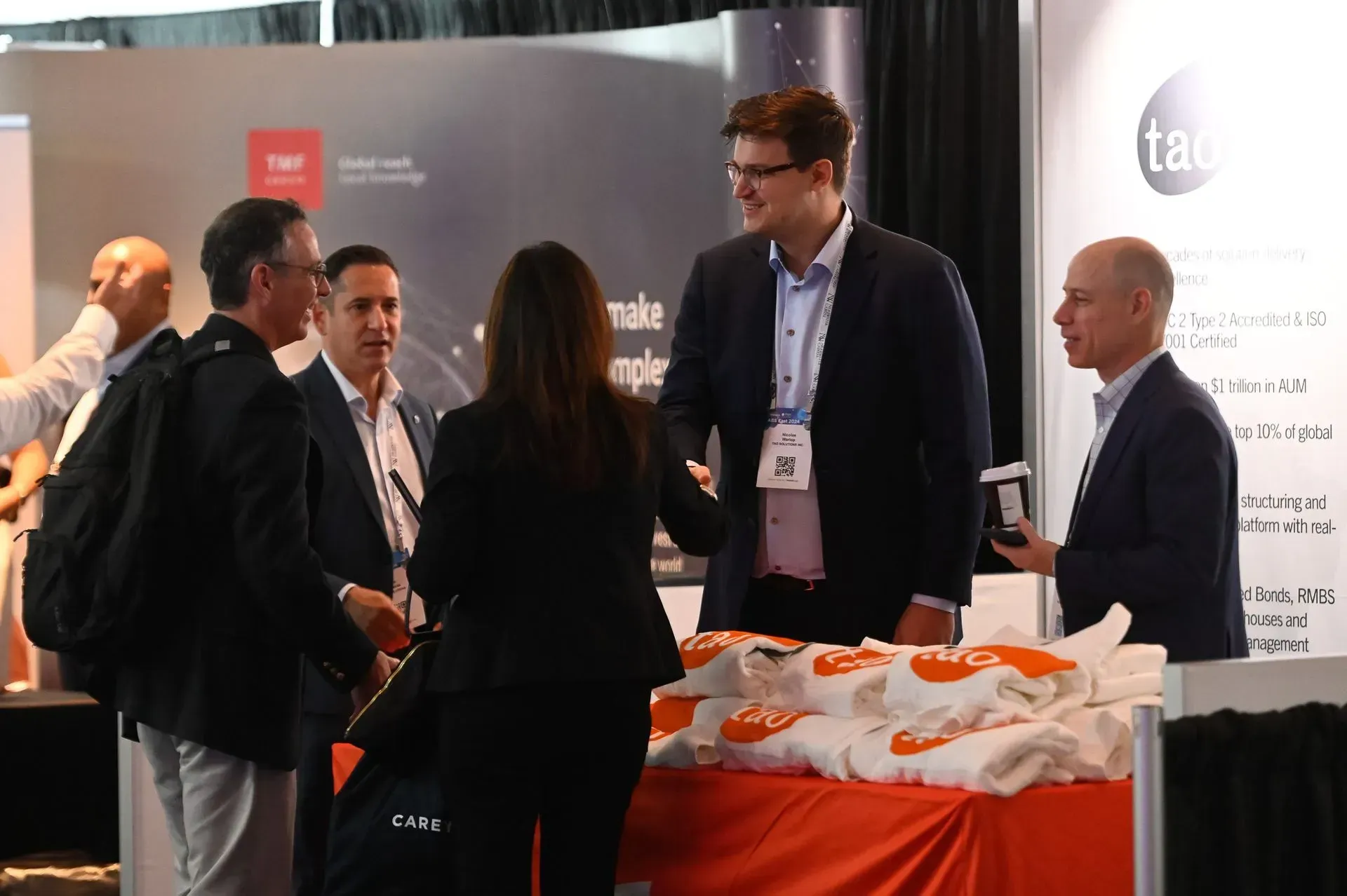 Five business professionals in suits converse at a trade show booth with orange branded items on a table.