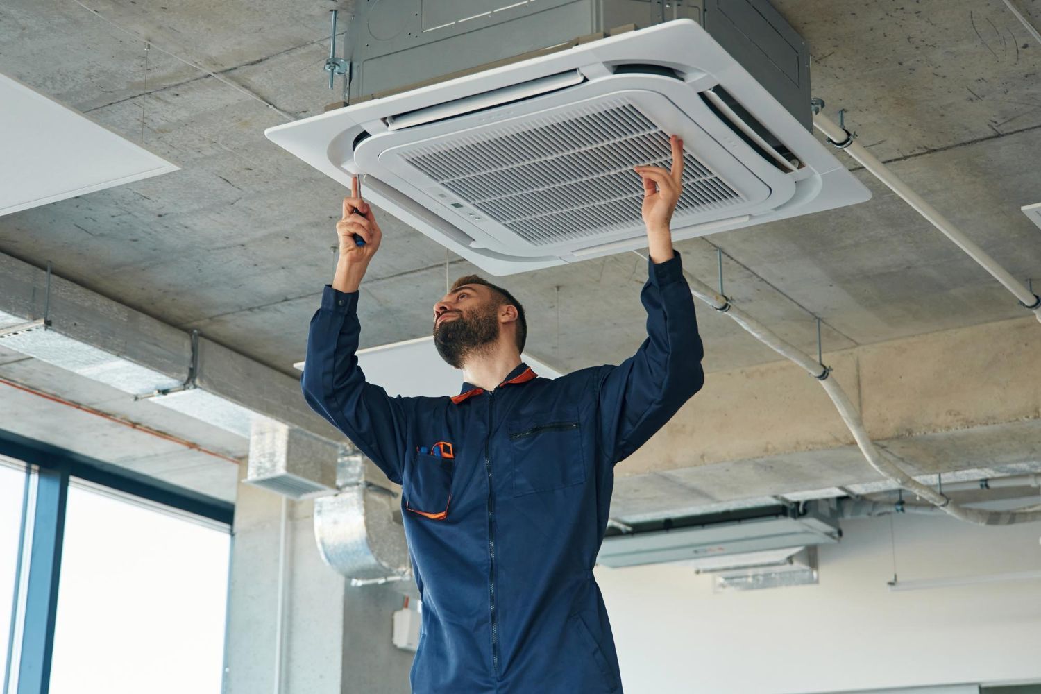 Technician installing a ceiling cassette air conditioner in a bright industrial room.