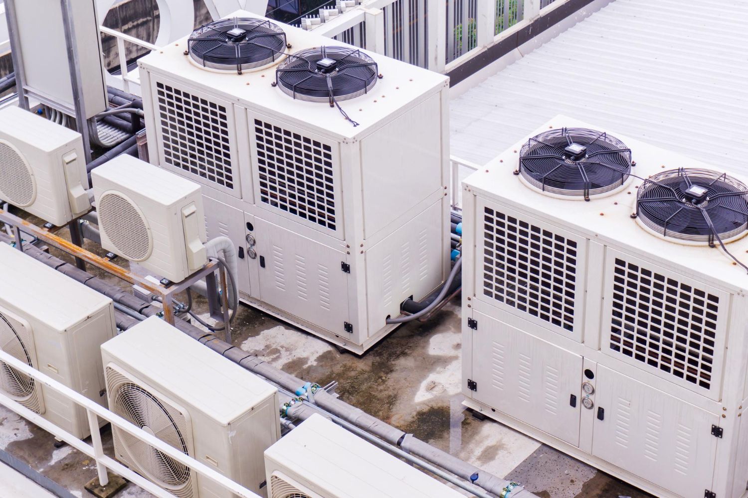 Rooftop HVAC units with white enclosures and fans beside building windows and pipes