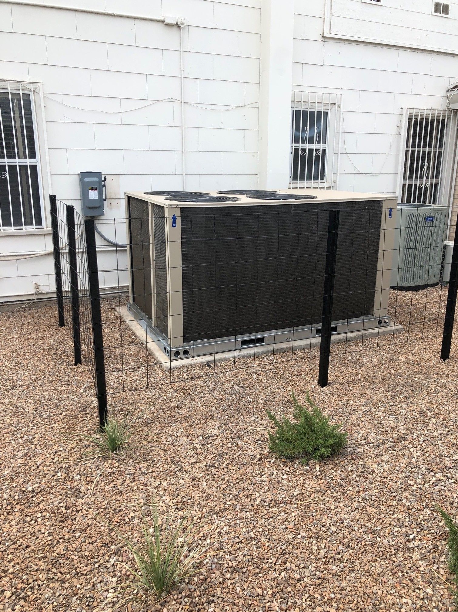 Outdoor HVAC unit beside a white house, surrounded by gravel and black metal fencing.